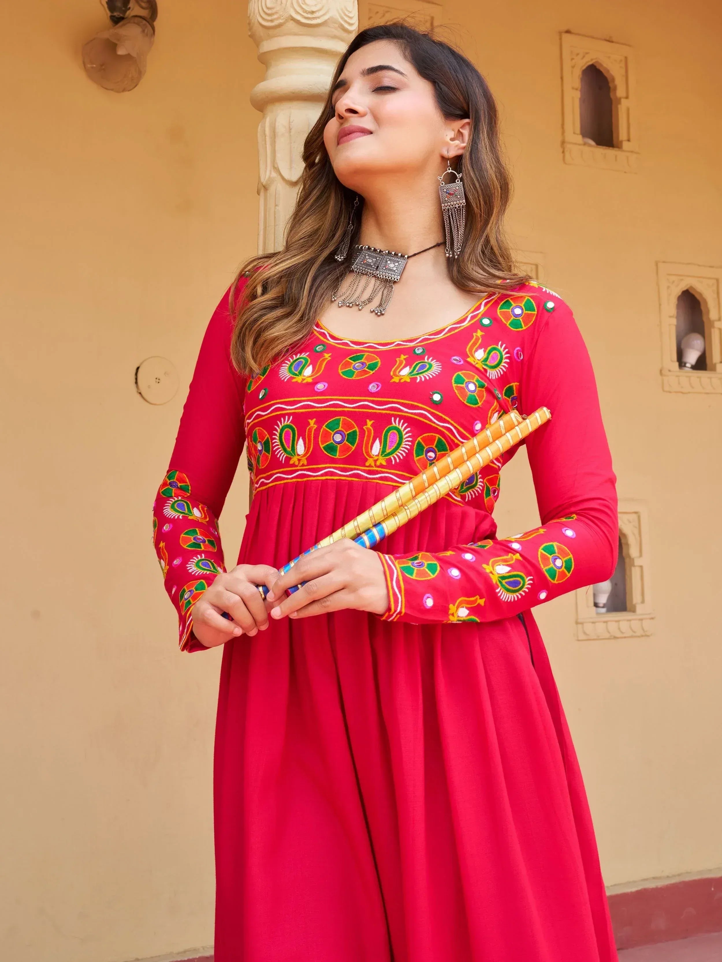 Woman in red embroidered ethnic gown with peacock motifs, holding dandiya sticks, traditional Indian wear