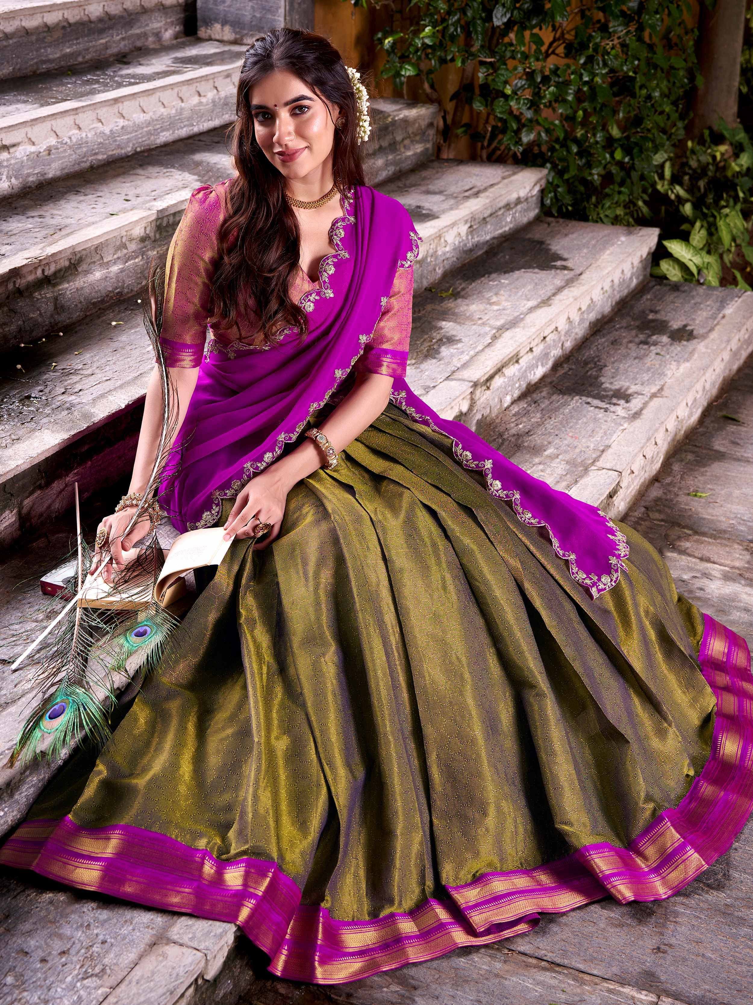 Woman in purple and olive green embroidered lehenga choli, sitting on stone steps with peacock feathers