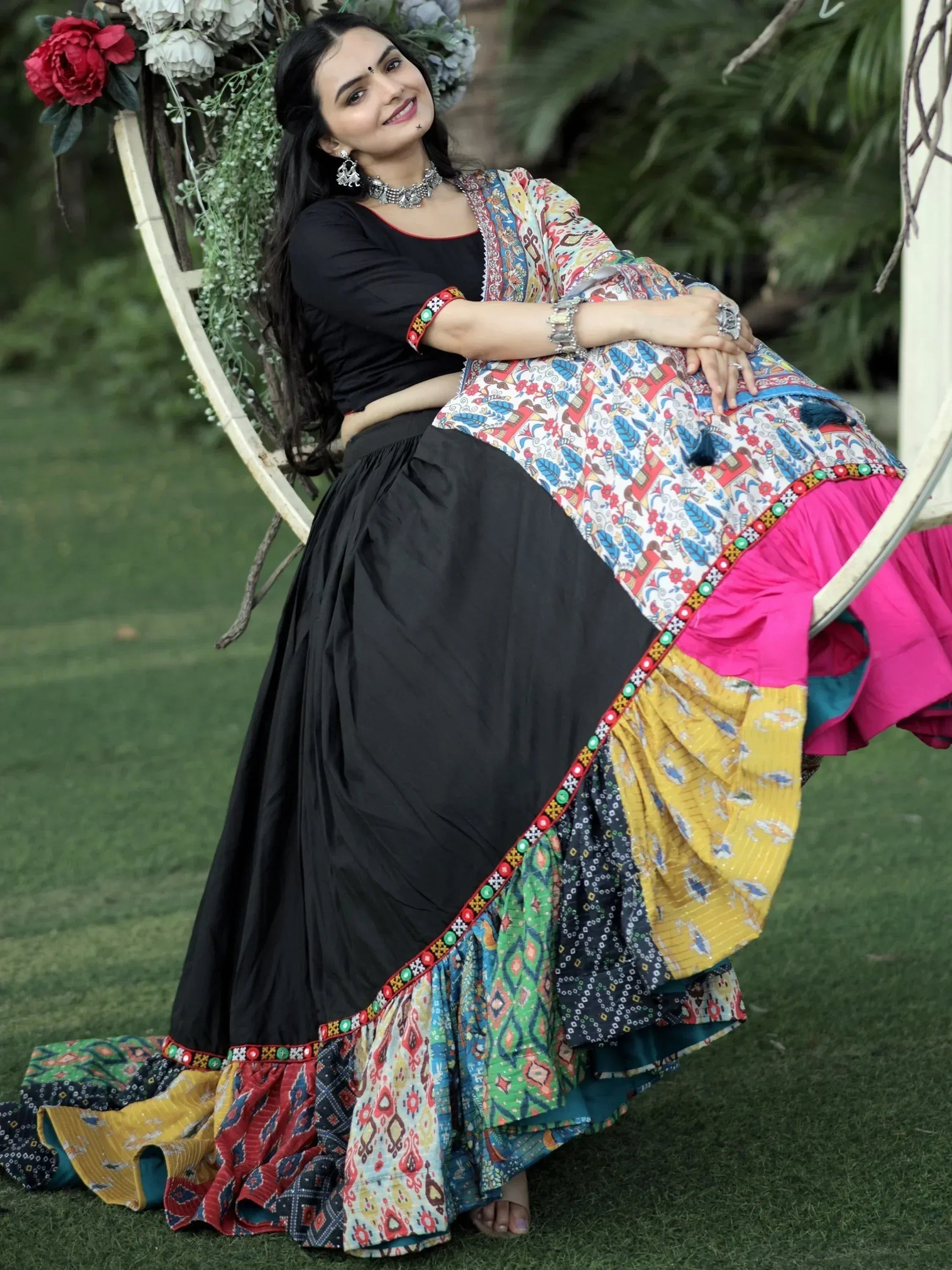 Woman in colorful Indian ethnic lehenga choli with black top, floral dupatta, and layered skirt, seated outdoors.