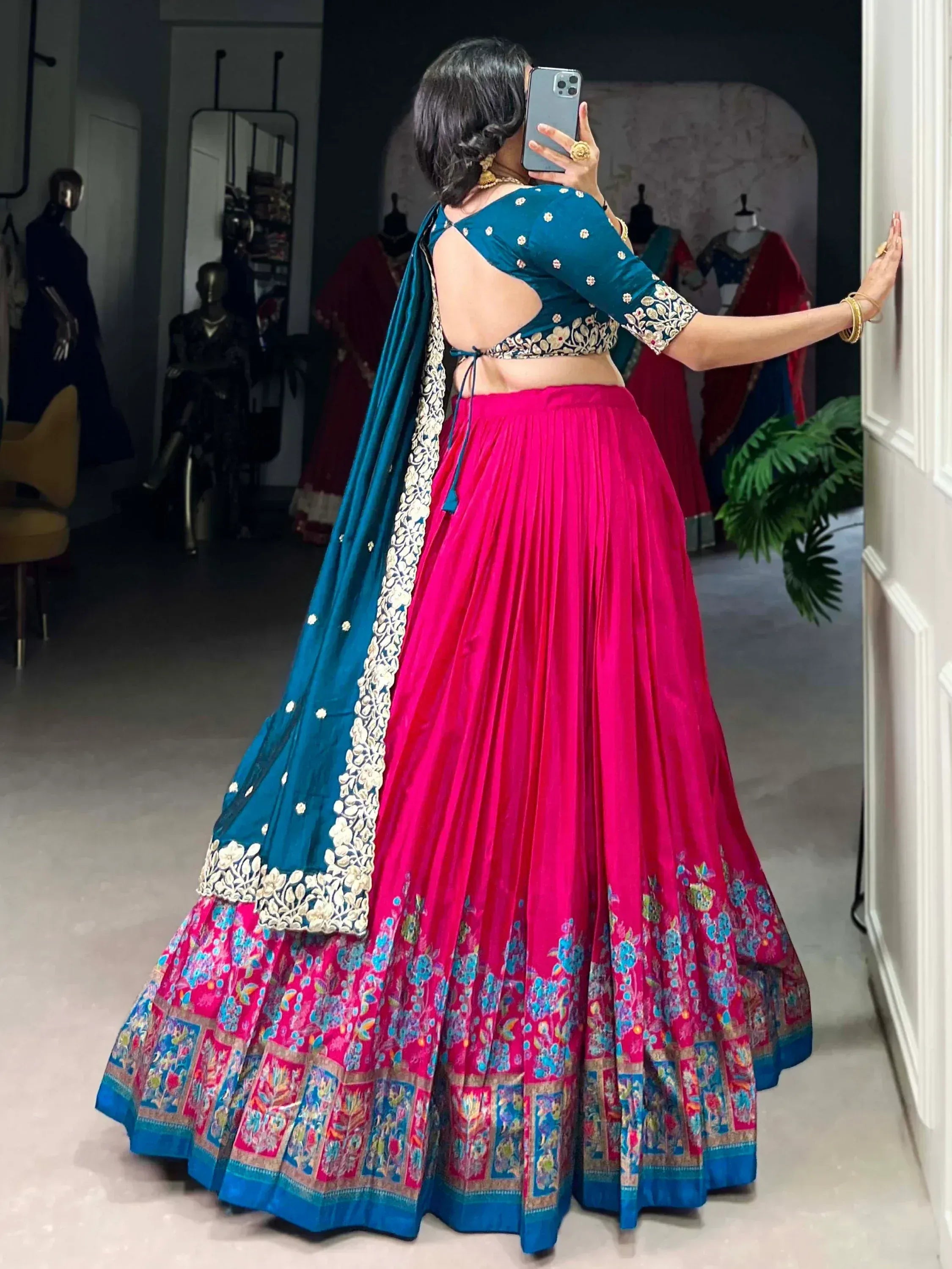 Woman in vibrant pink and teal designer lehenga choli with floral embroidery, taking mirror selfie in boutique