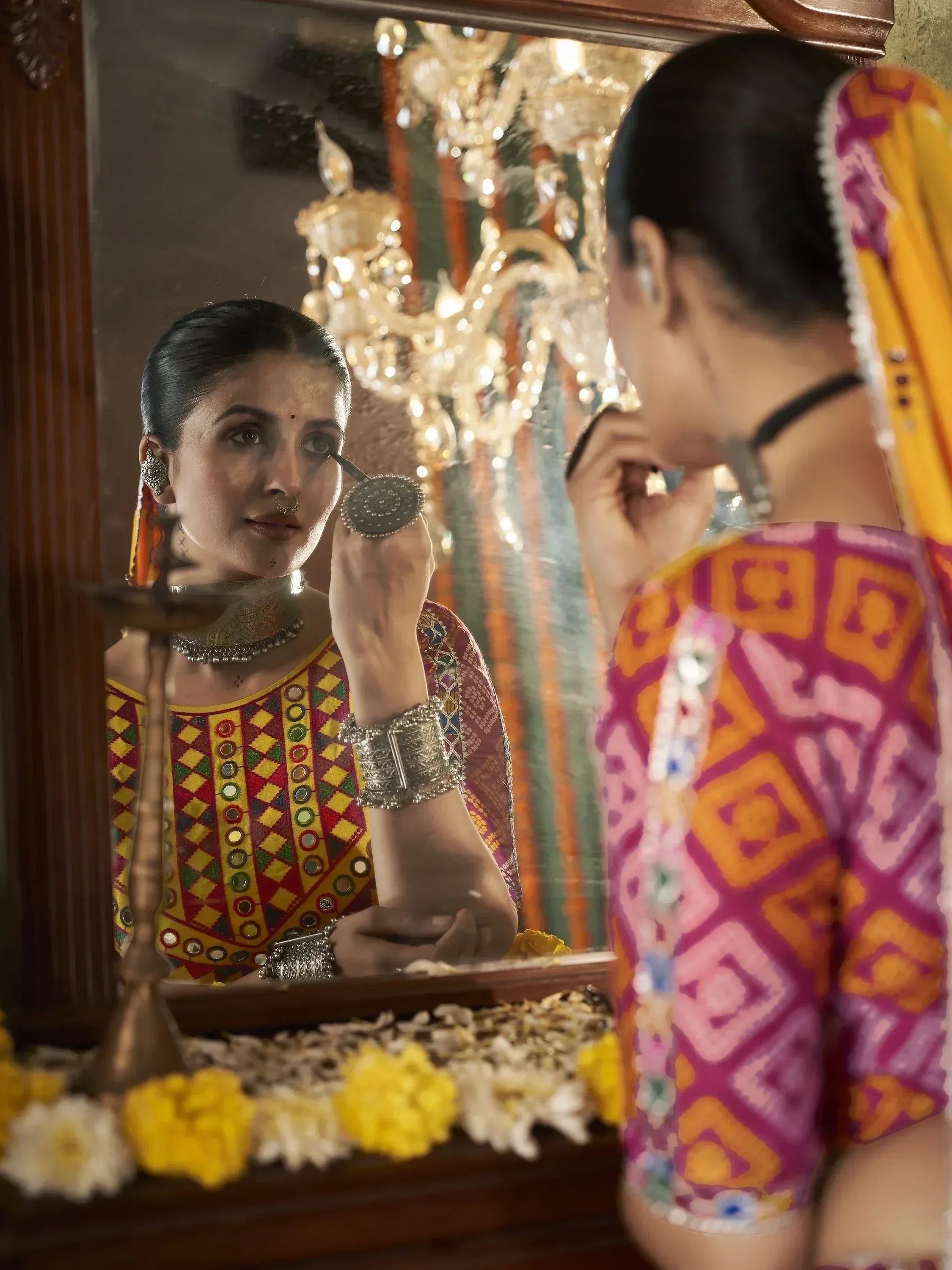 Woman in colorful ethnic wear applying makeup, reflected in mirror with chandelier, floral decor