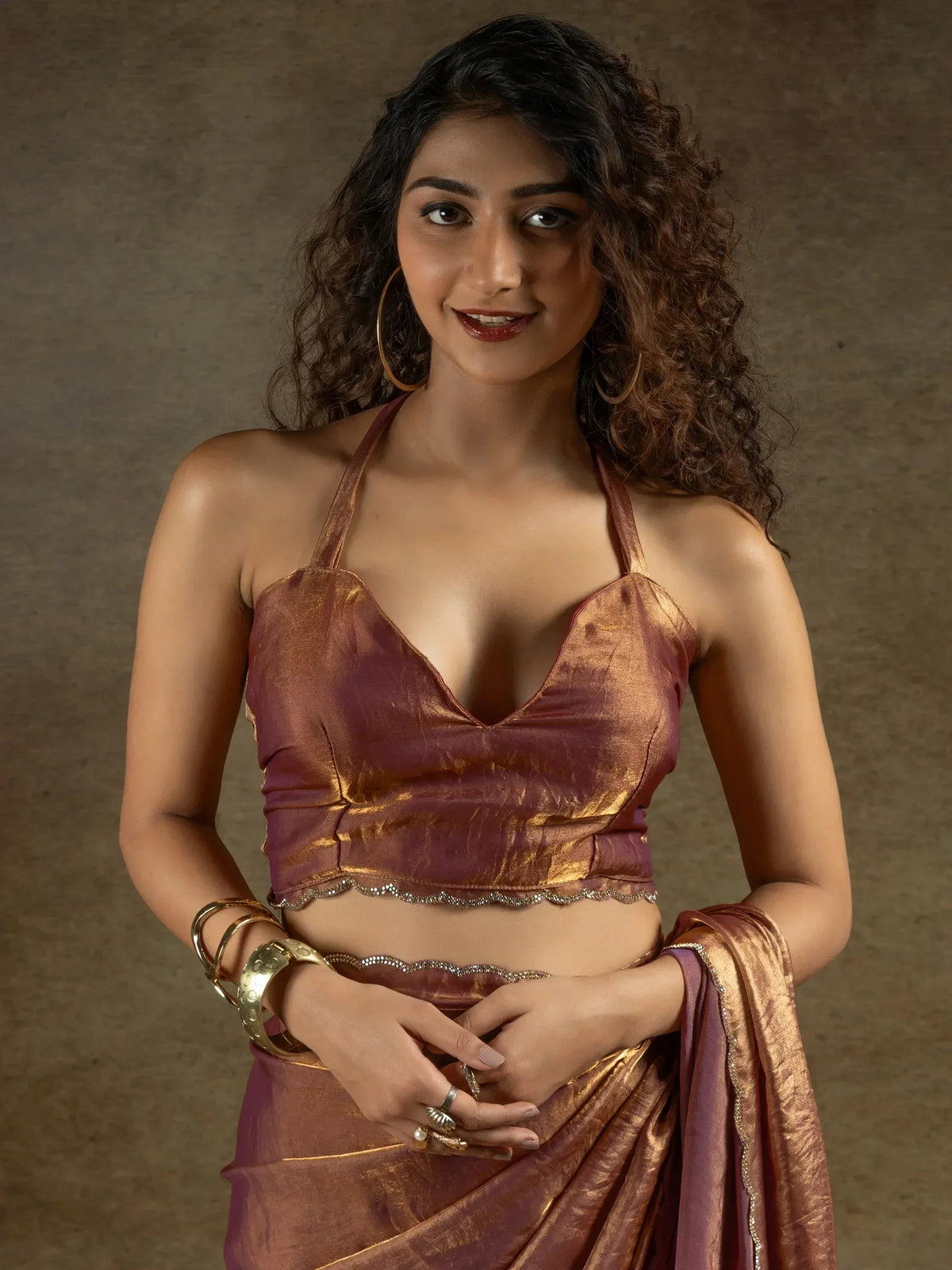 Woman in shimmering maroon and gold saree with halter neck blouse, ethnic jewelry, curly hair