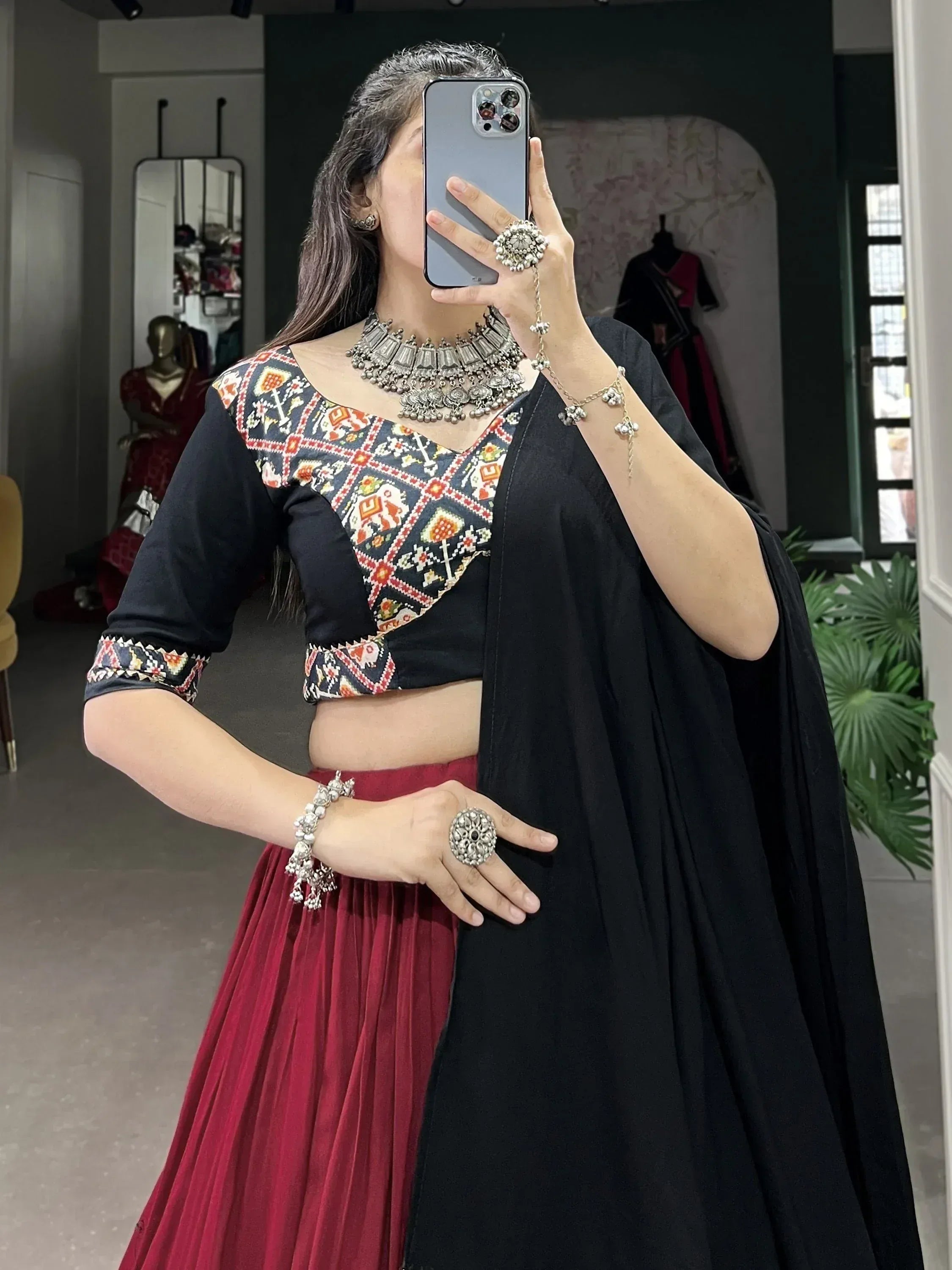 Woman in black embroidered crop top, maroon pleated skirt, and silver jewelry, taking mirror selfie in boutique