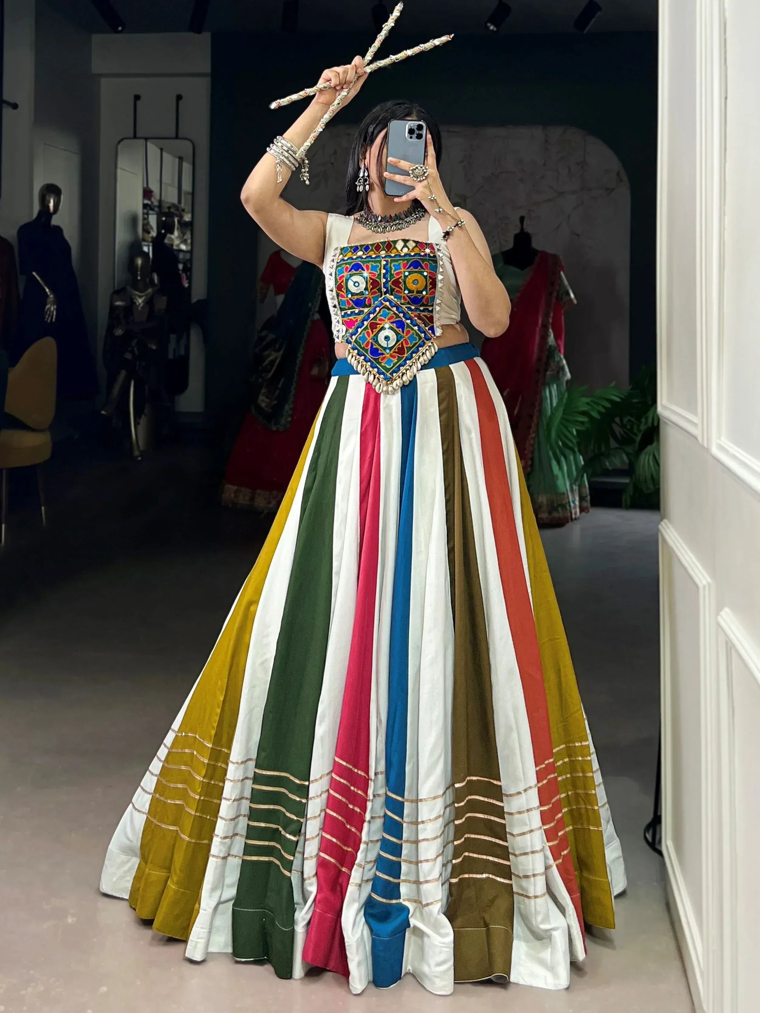 Woman in colorful striped lehenga with embroidered top, holding decorative sticks, perfect for festive Indian ethnic wear.