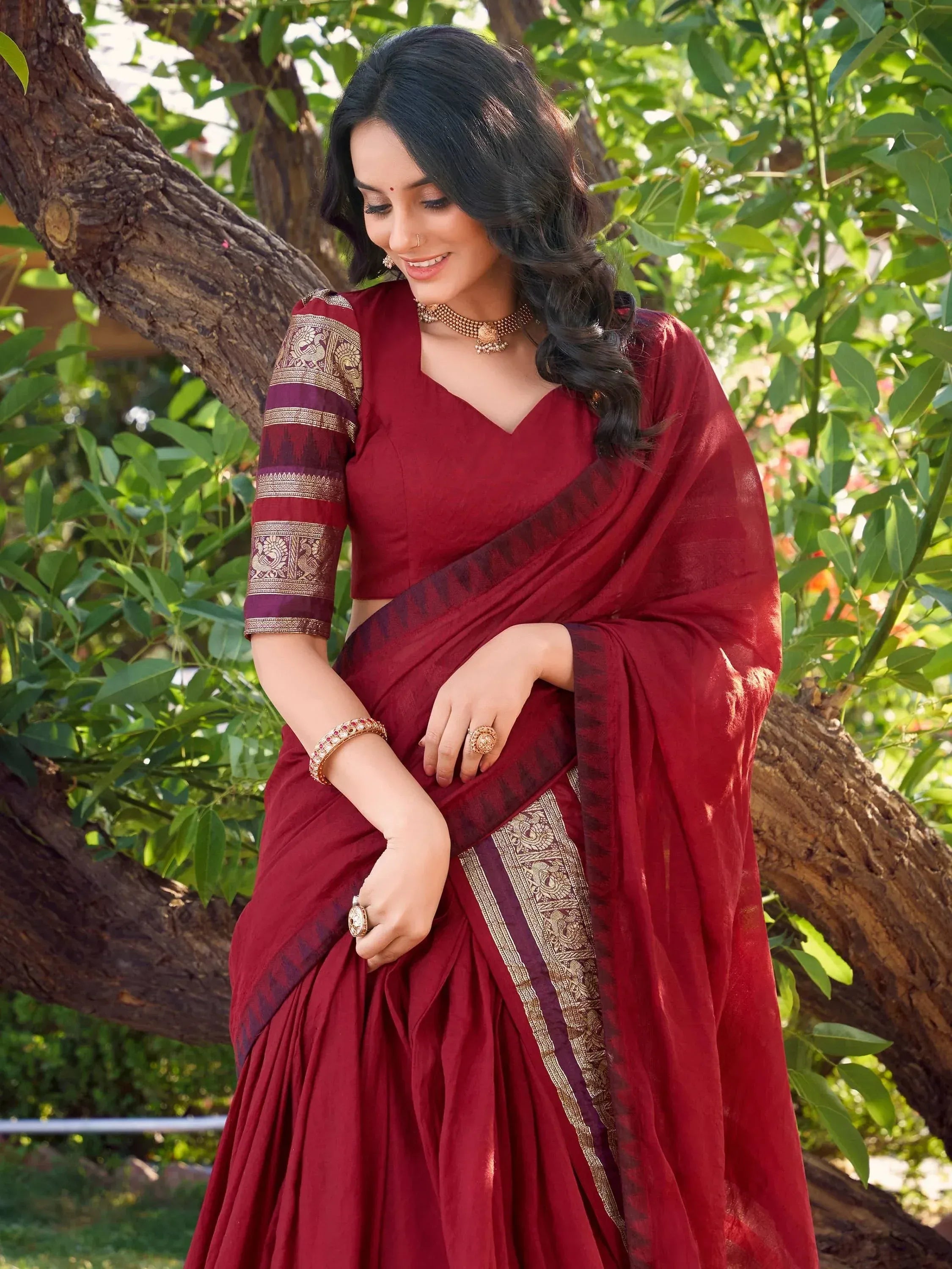 Woman in maroon ethnic saree with gold border, styled for festive occasions under green trees