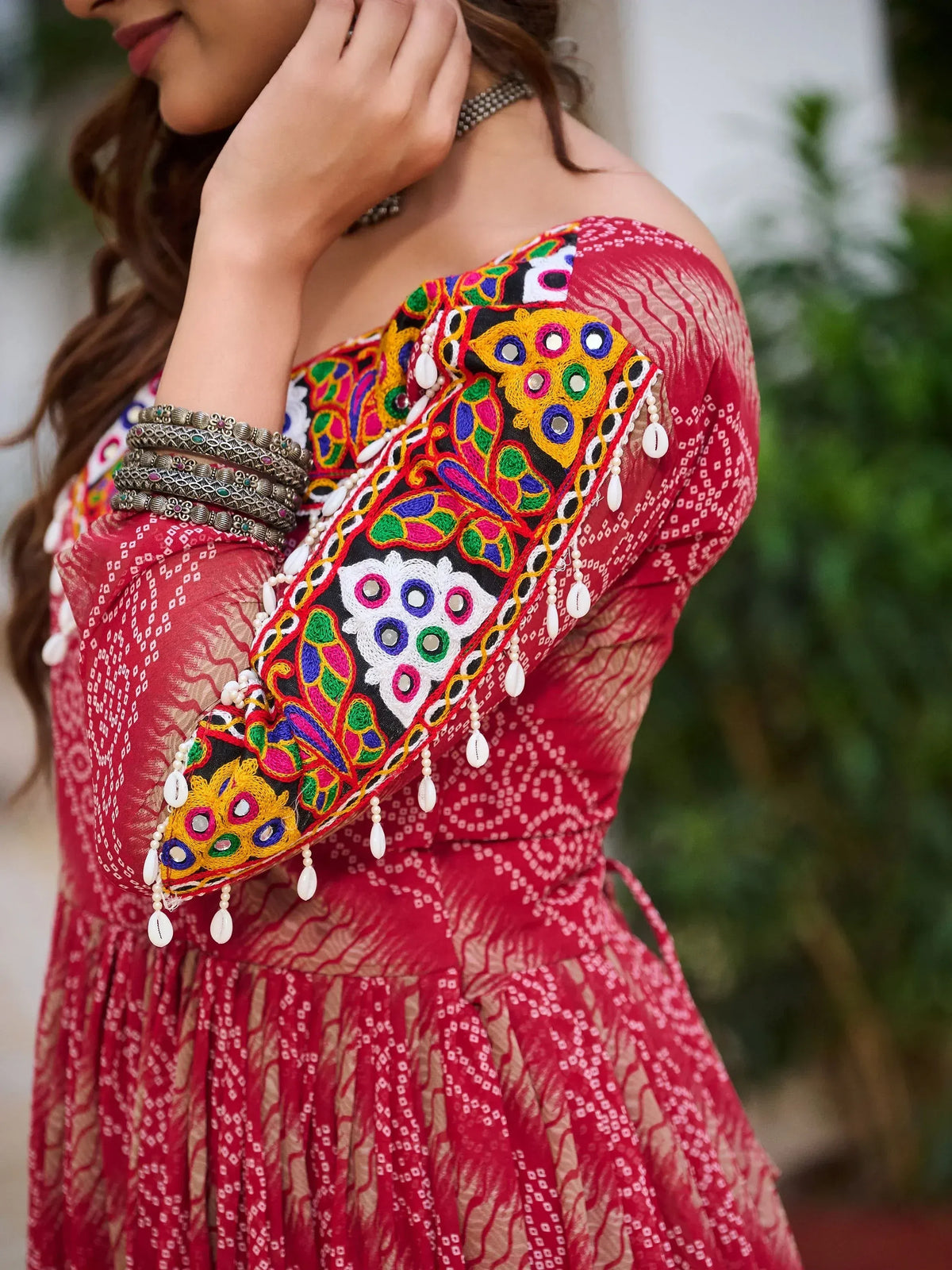 Red off-shoulder ethnic gown with colorful mirror embroidery and shell tassels, perfect for Indian festive wear.
