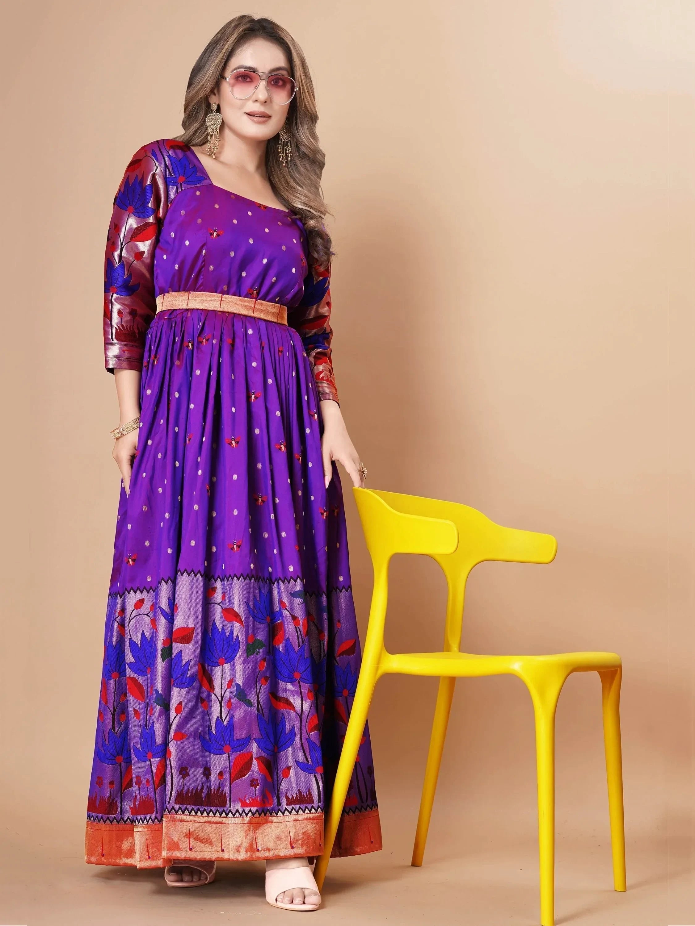 Woman in a purple ethnic gown with floral patterns and gold accents, posing by a yellow chair