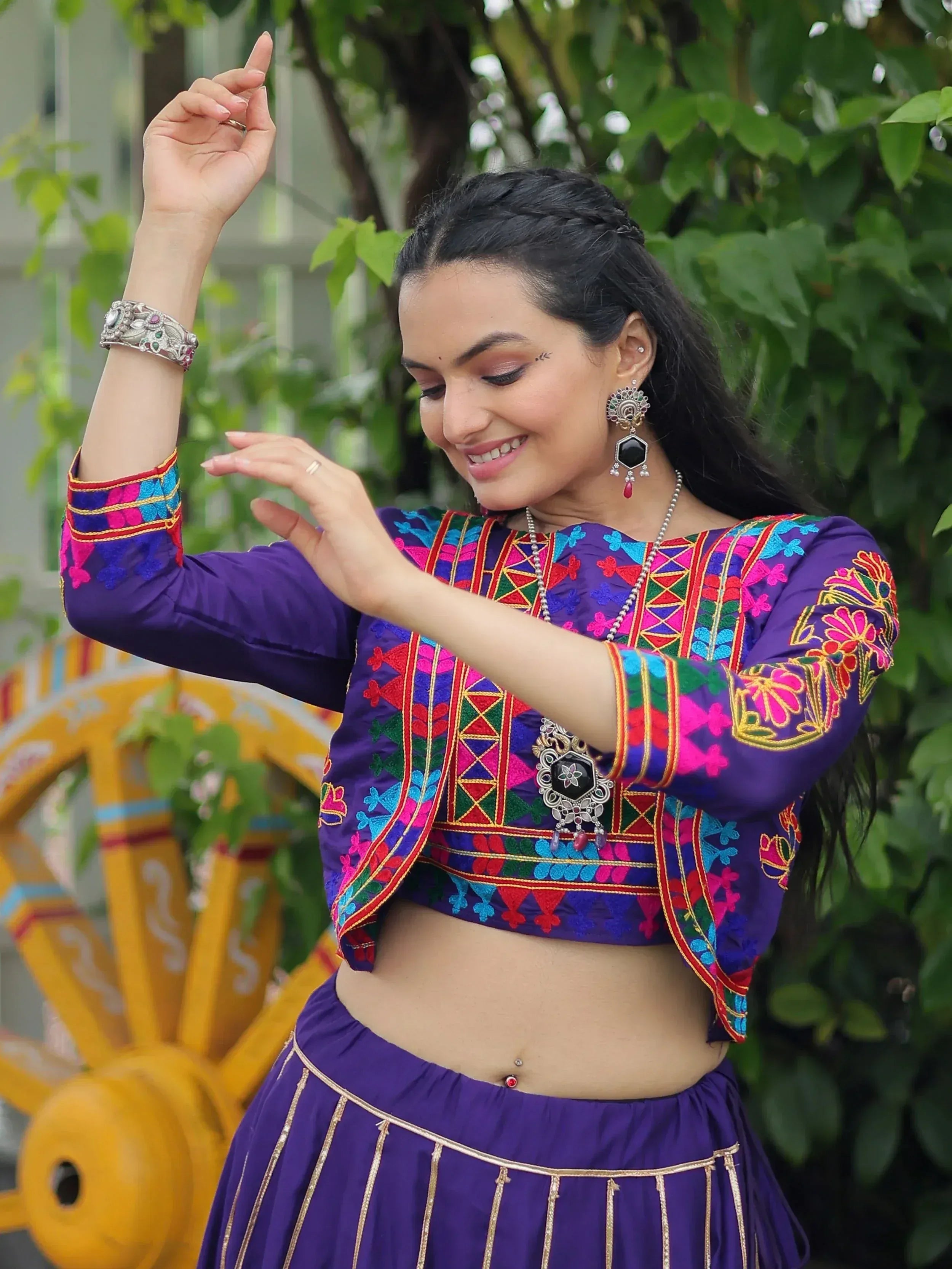Woman in vibrant purple embroidered ethnic top and skirt, dancing outdoors with traditional jewelry