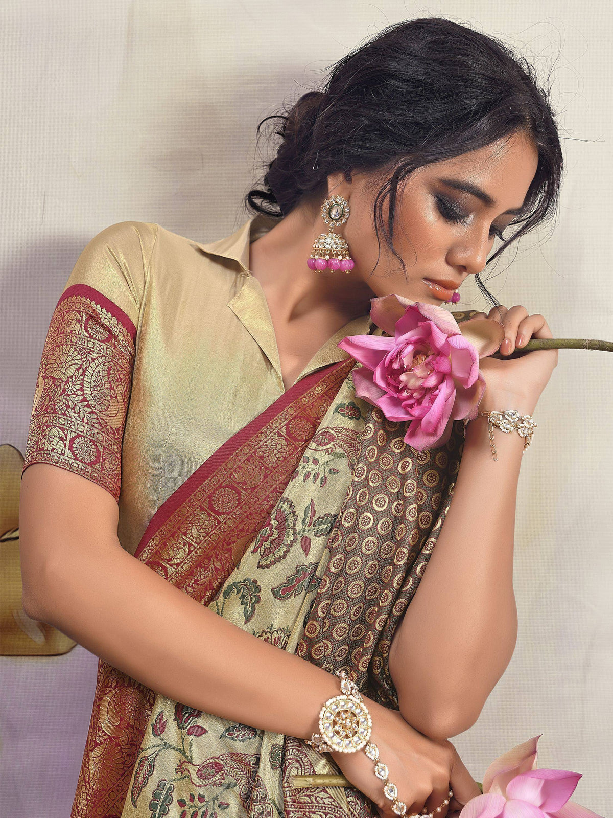 Woman in elegant gold and maroon ethnic saree with floral print, holding pink lotus, adorned with jhumka earrings and traditional jewelry.