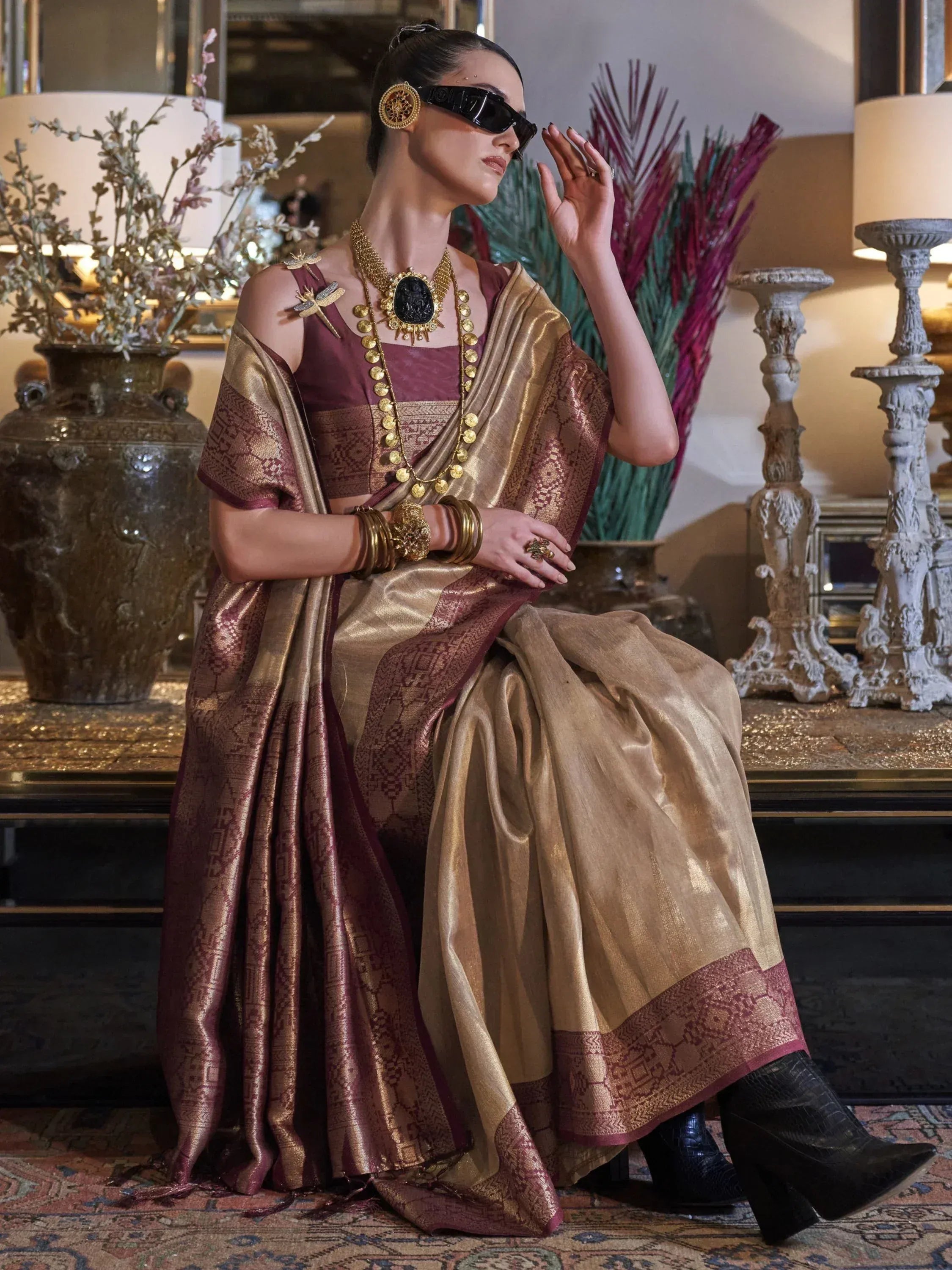 Woman in maroon and gold designer saree with heavy jewelry, modern sunglasses, and boots for ethnic fashion