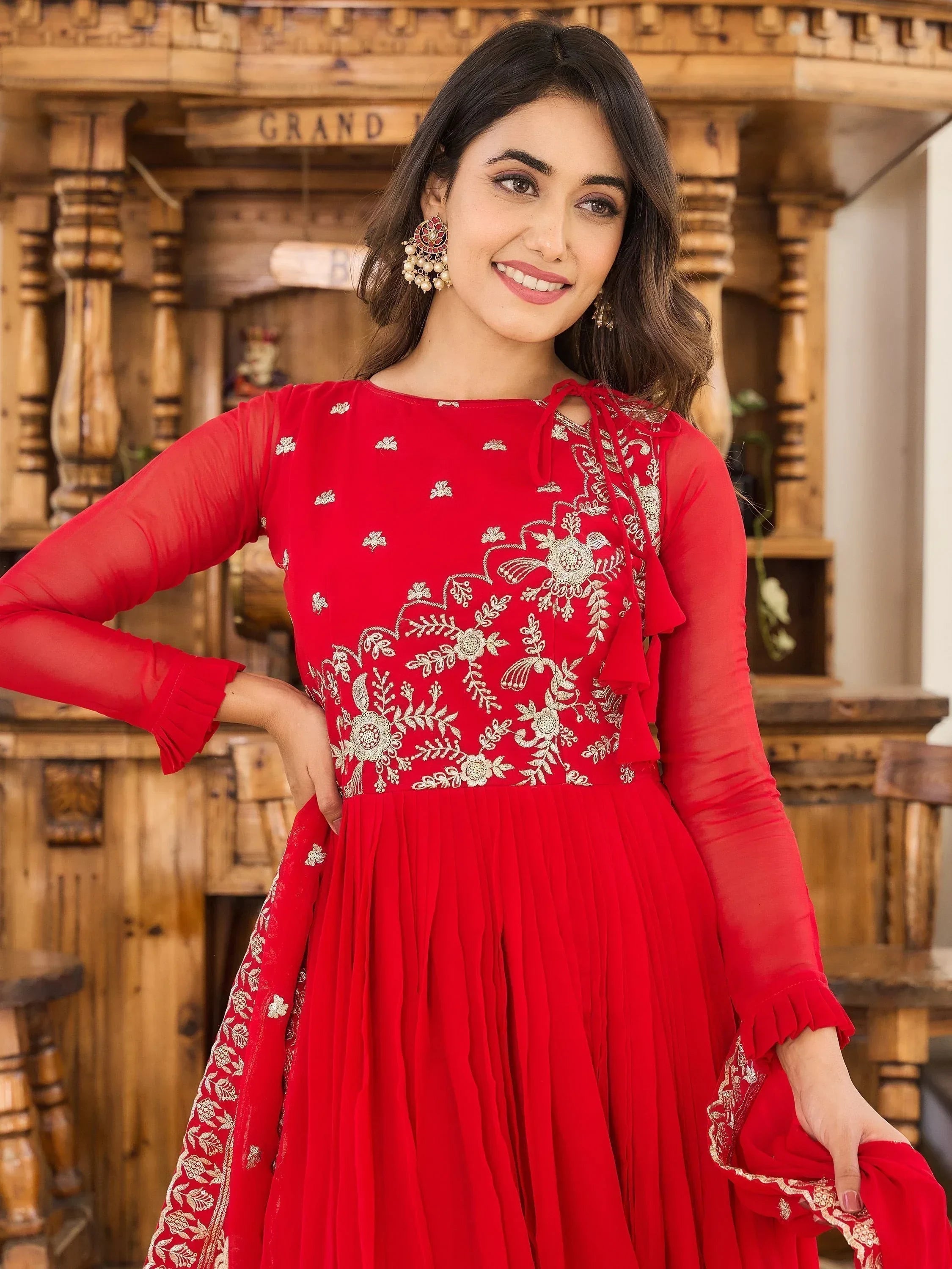 Woman in red embroidered ethnic gown with sheer sleeves and floral patterns, smiling indoors