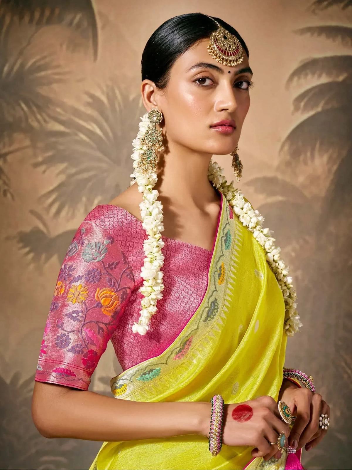 Woman in vibrant yellow saree with pink floral blouse, traditional Indian jewelry, and jasmine garland