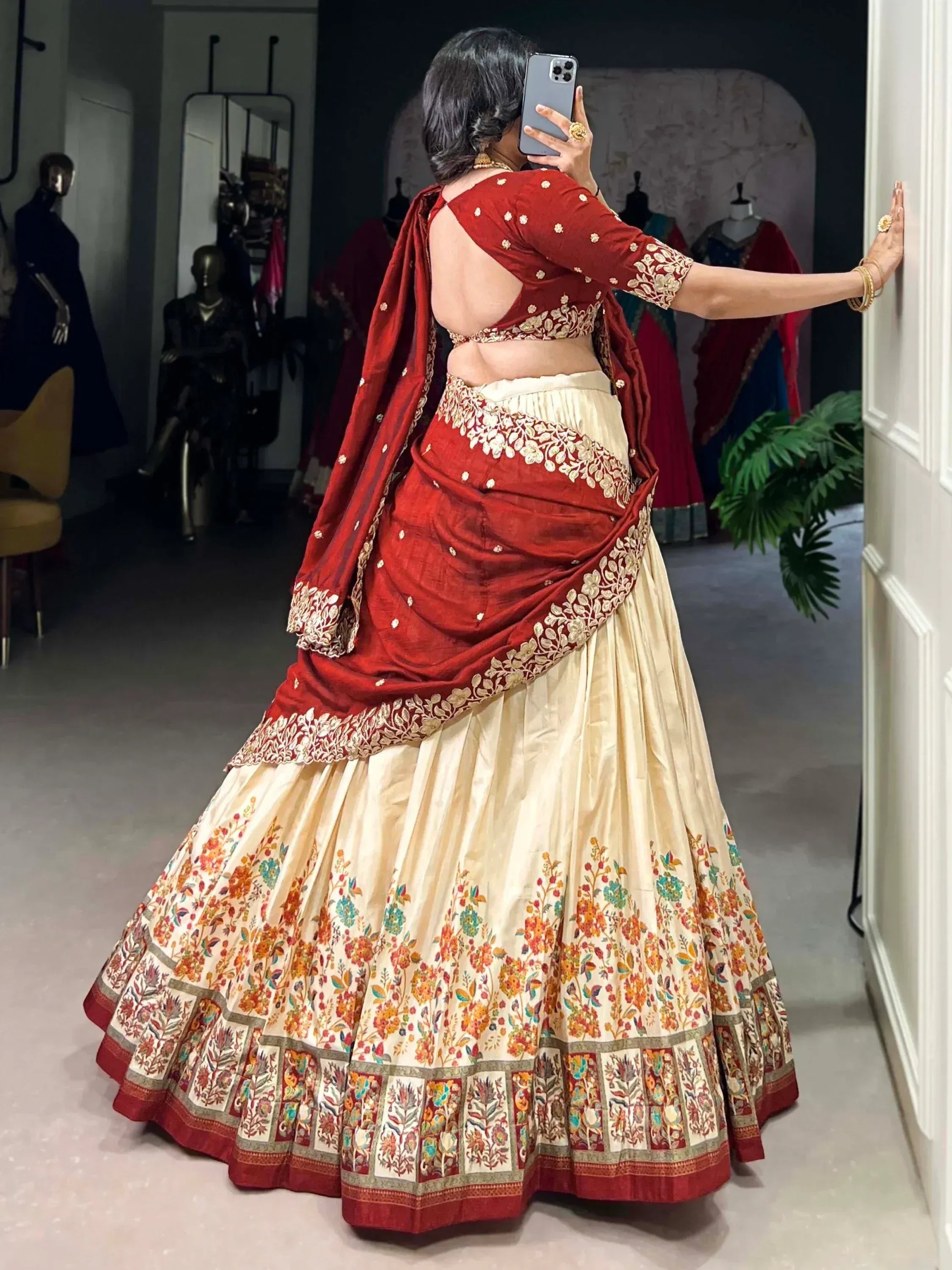 Woman in red and cream floral lehenga choli with backless blouse, taking mirror selfie in boutique
