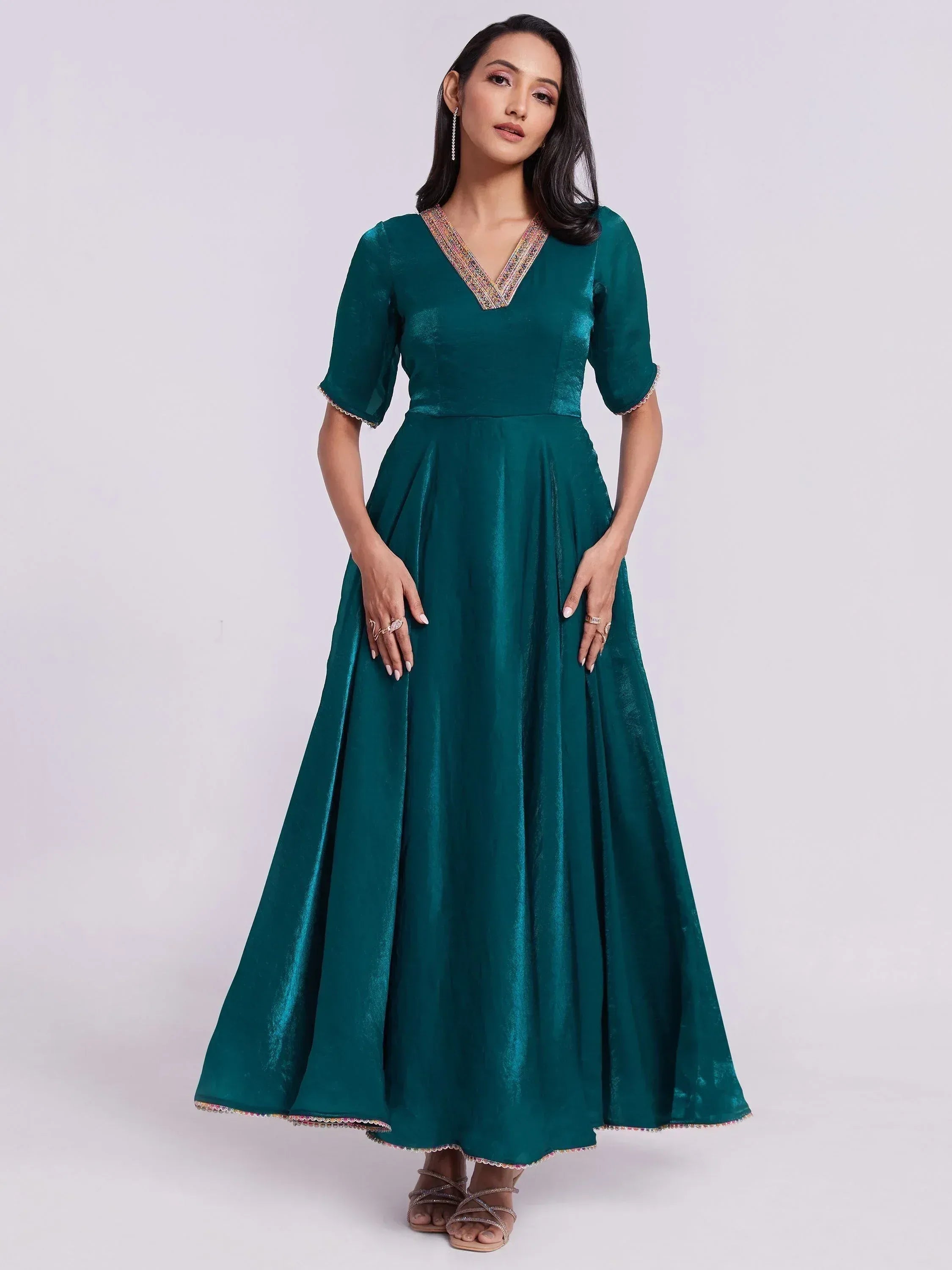 Woman wearing a teal silk ethnic gown with V-neck and half sleeves, standing against plain background.