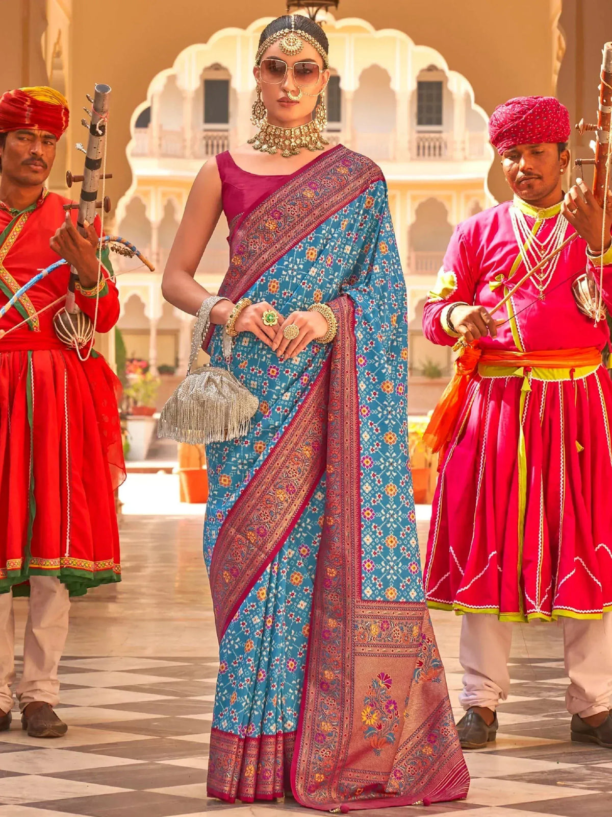 Woman in blue floral saree with pink border, traditional Indian jewelry, flanked by musicians in red attire