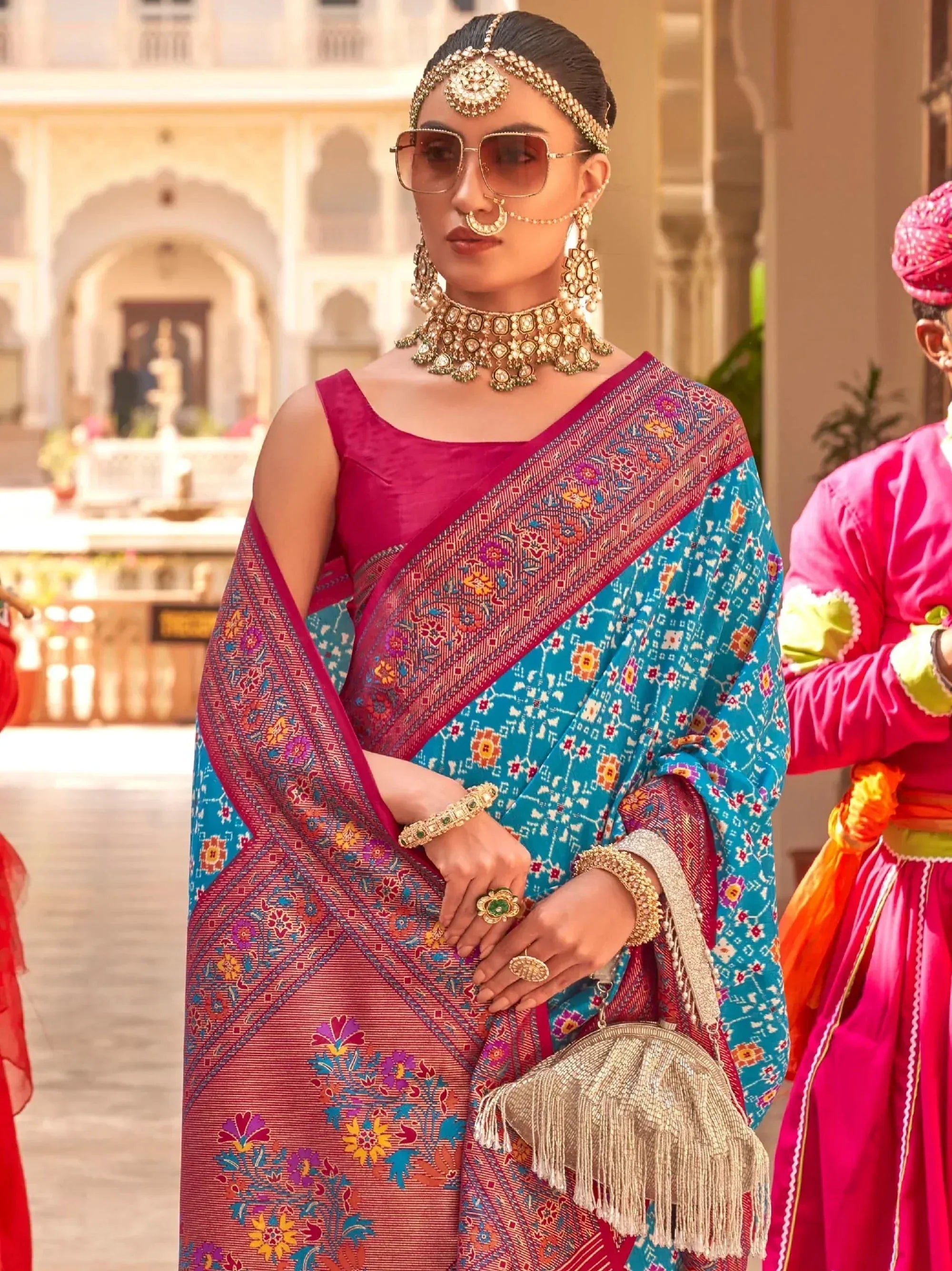 Woman in vibrant blue and pink floral saree with traditional Indian jewelry and sunglasses, styled for festive occasions.
