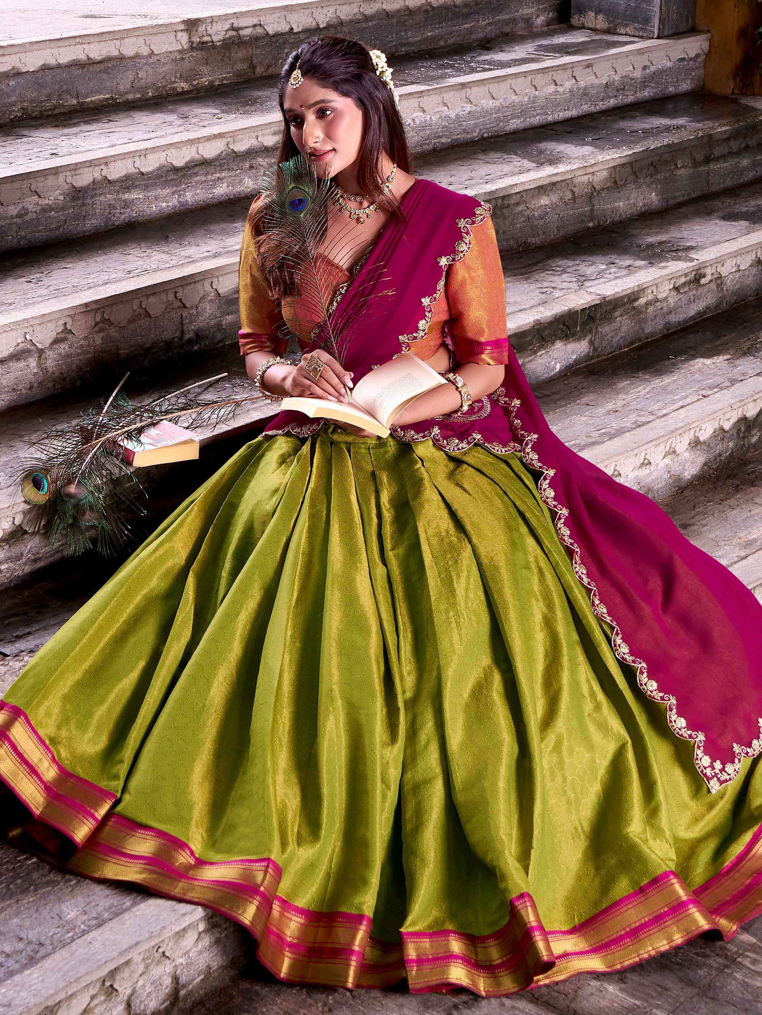 Woman in vibrant green and magenta lehenga choli with gold embroidery, sitting on stone steps holding a book and peacock feather.