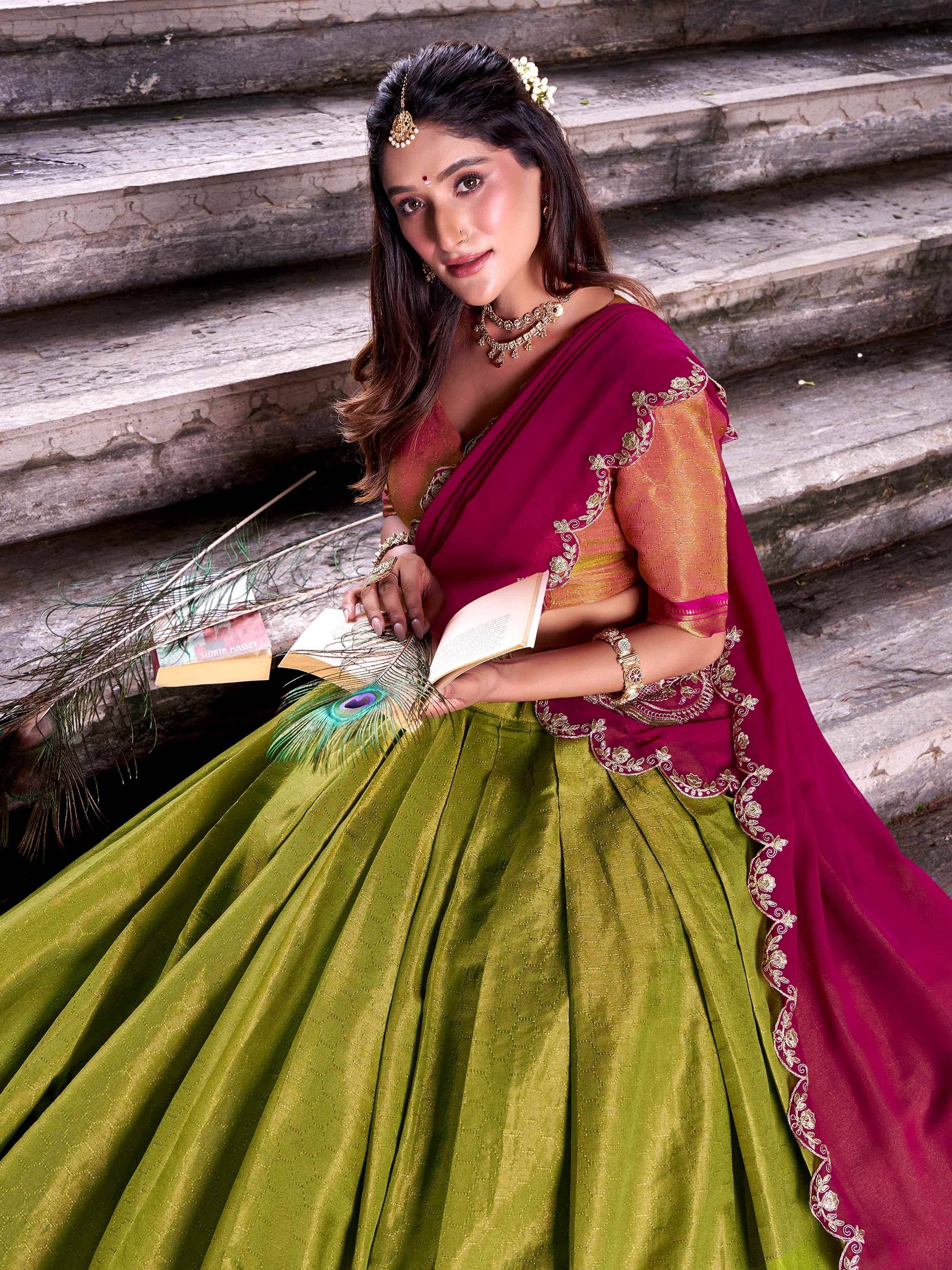 Woman in maroon and green designer lehenga choli with gold embroidery, holding book and peacock feather on stone steps