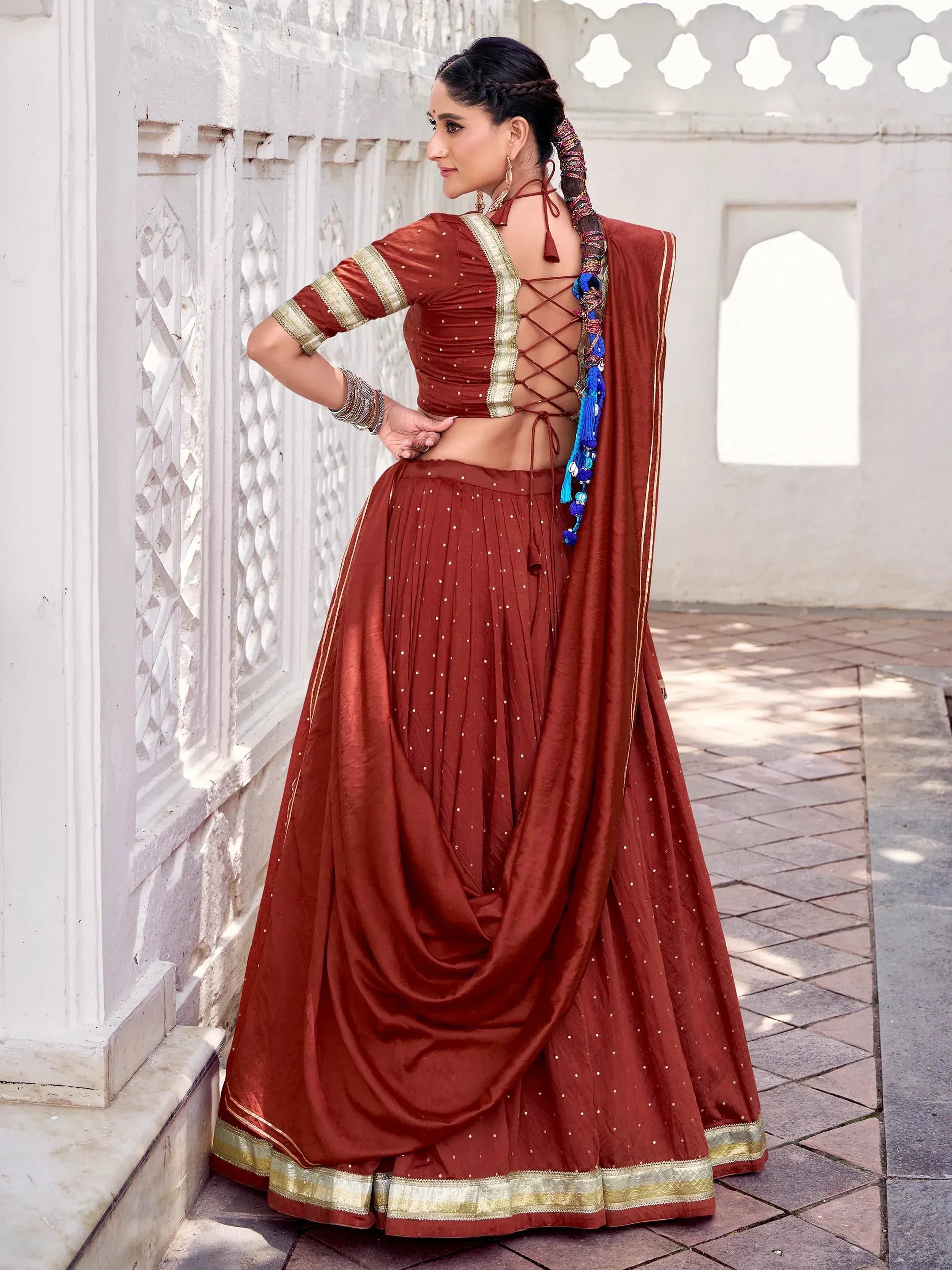 Rust Indian lehenga choli with back lace-up blouse, gold trim, and blue tassels, styled for ethnic wear