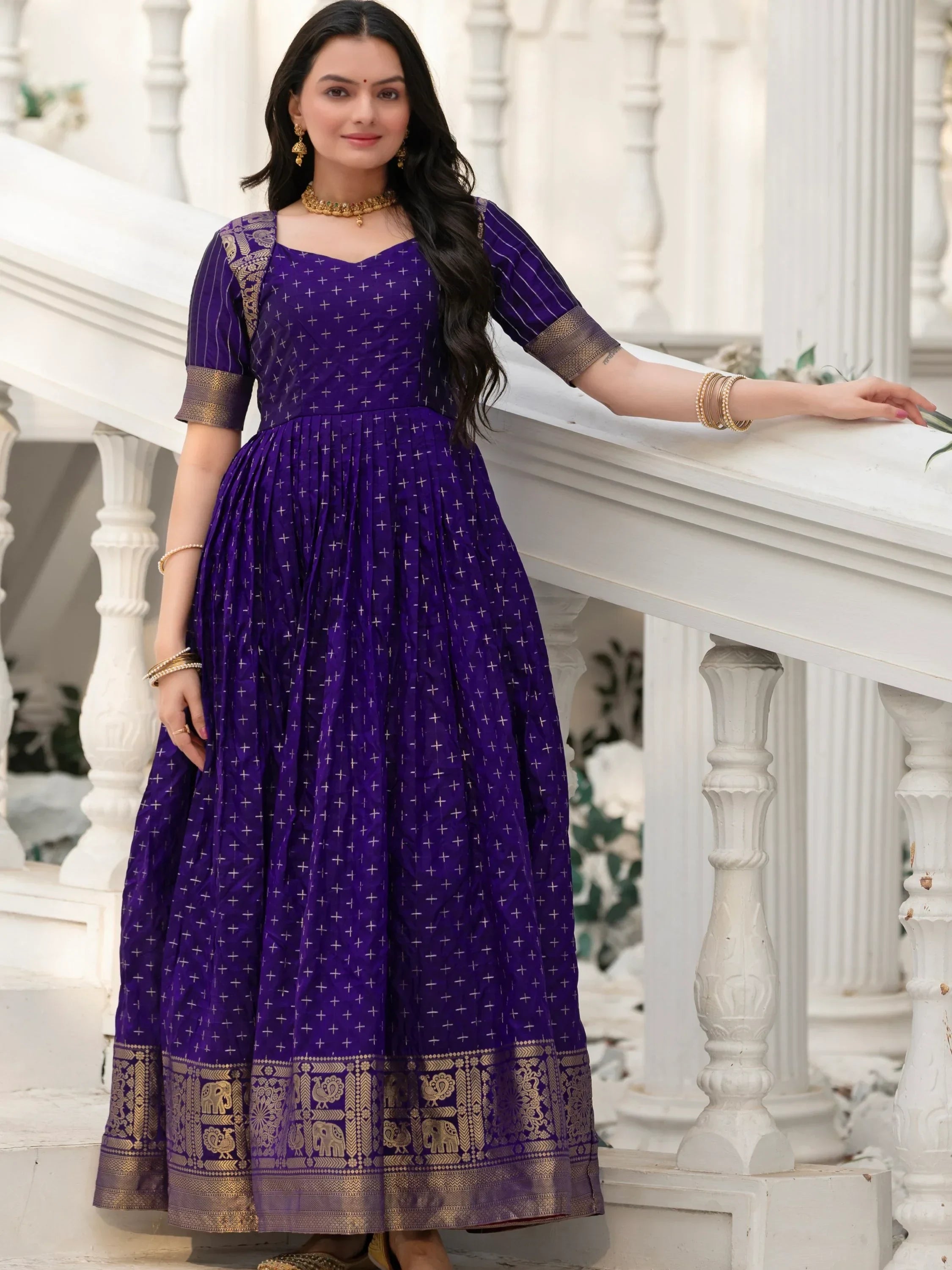 Woman in a purple ethnic gown with gold motifs and traditional jewelry, standing by white railing