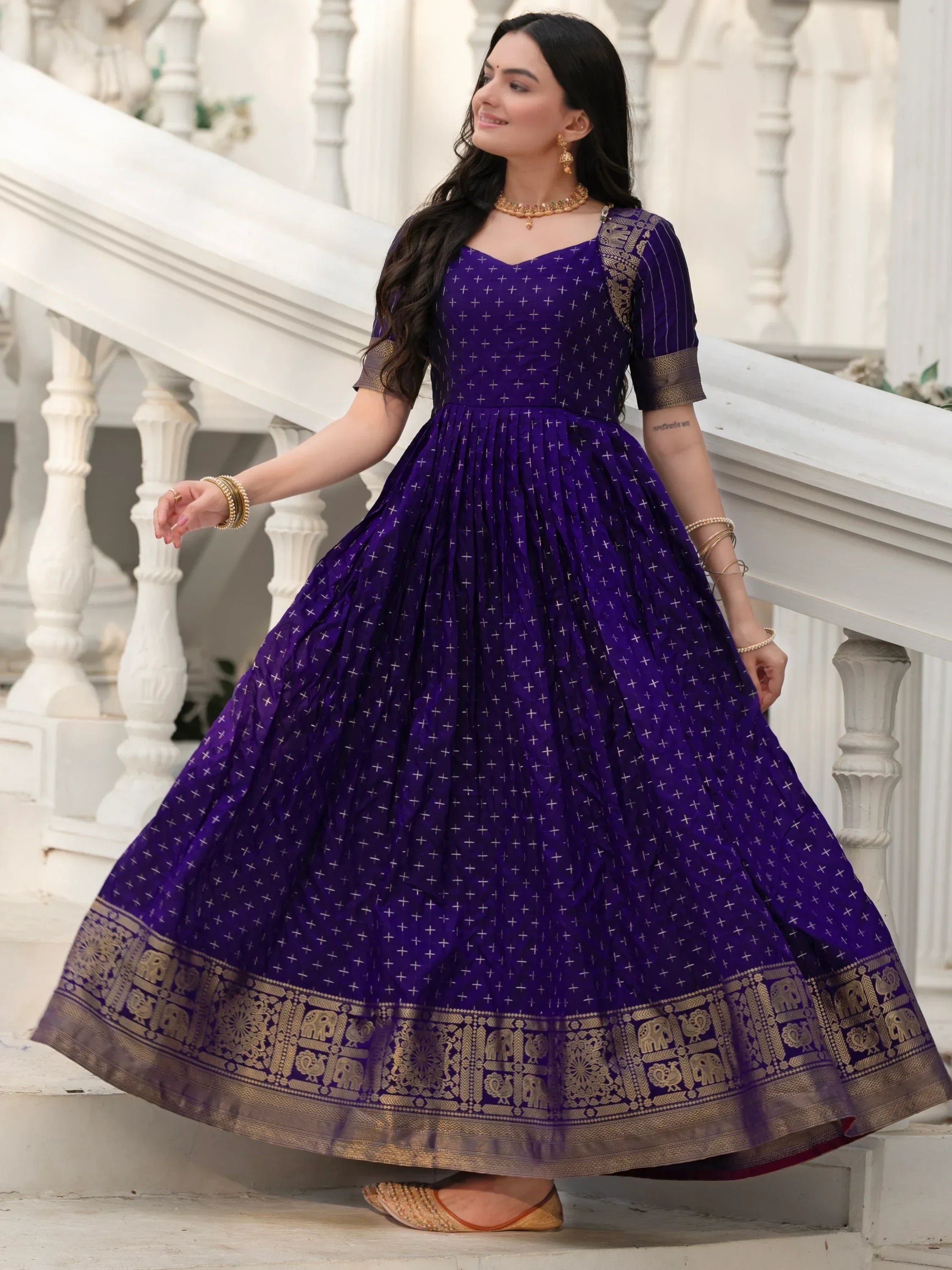 Woman in a purple ethnic gown with gold motifs and border, standing on white stairs