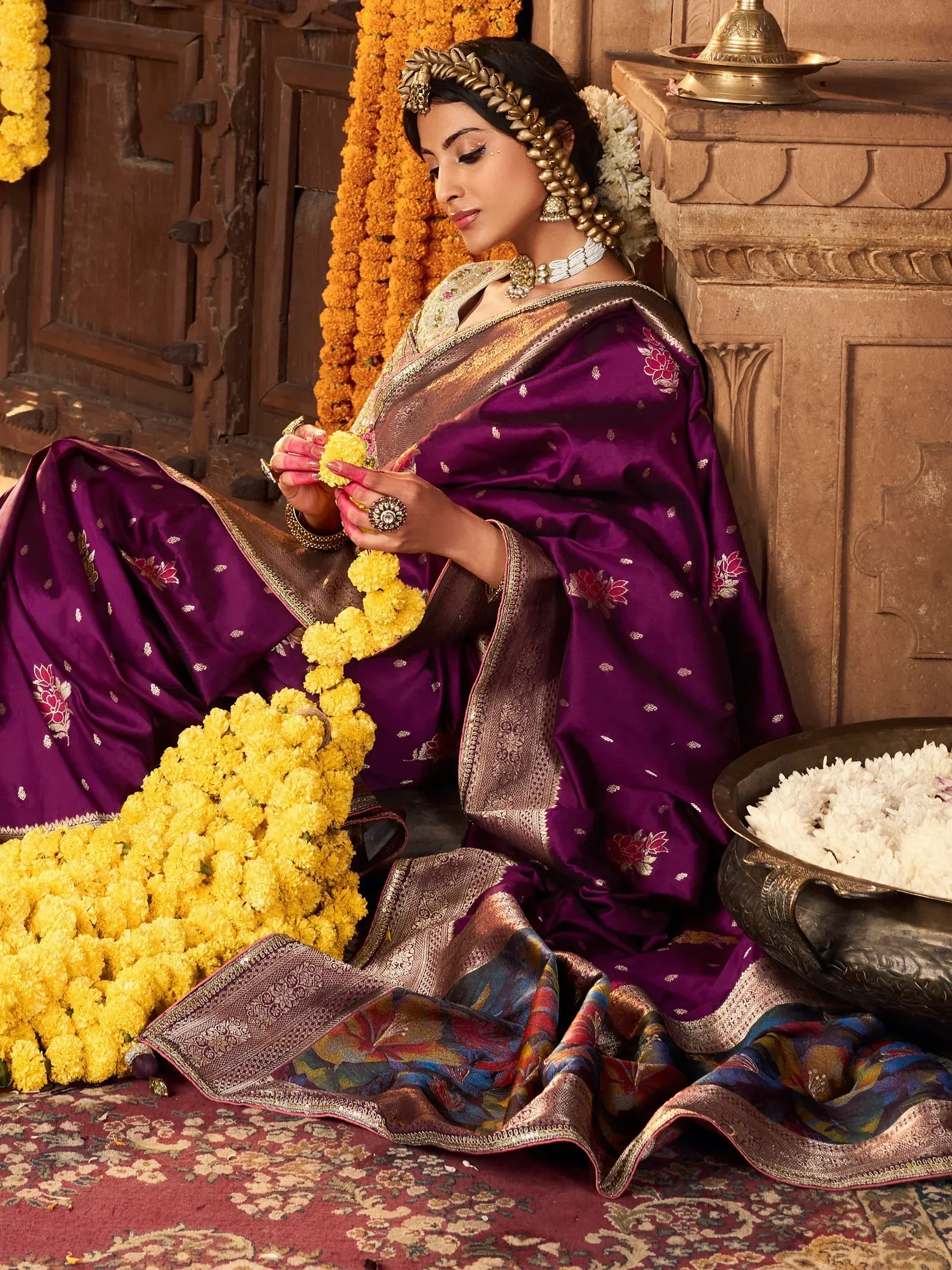 Woman in purple embroidered saree with gold jewelry, holding marigold garland, traditional Indian ethnic wear
