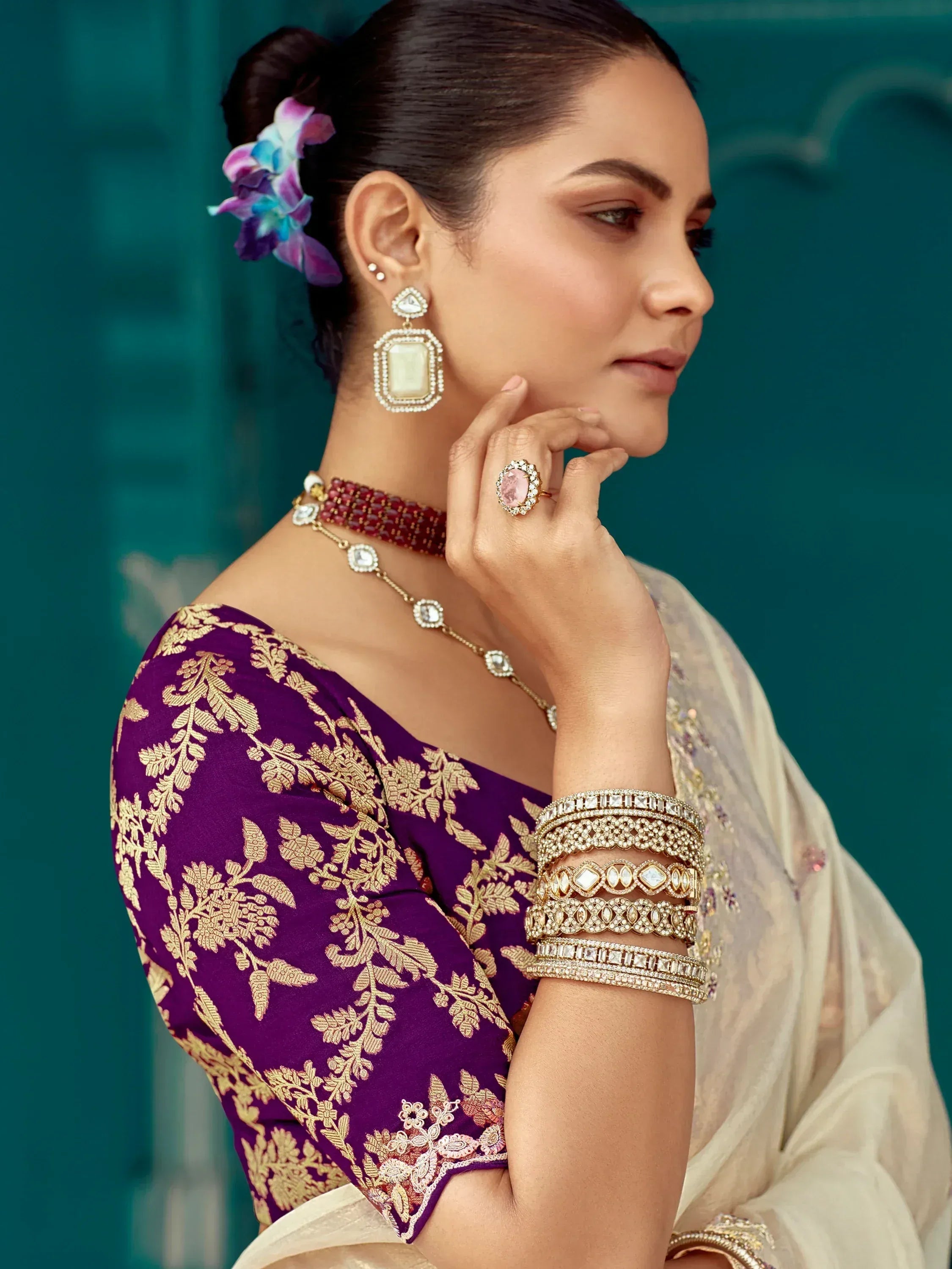 Woman in purple and gold embroidered saree with statement jewelry, floral hair accessory, and elegant pose