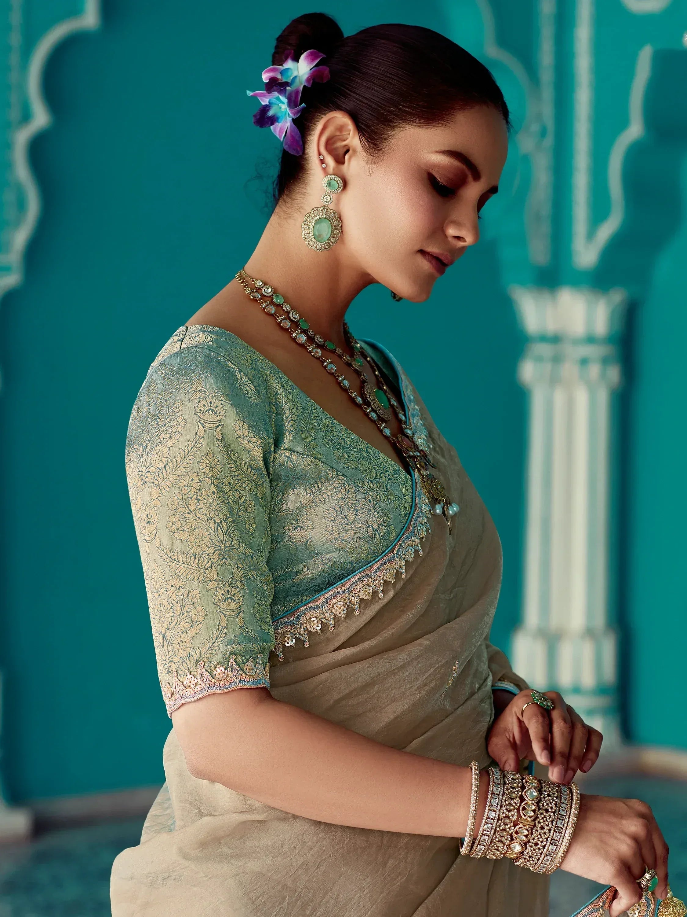 Elegant woman in designer saree with gold embroidery, statement jewelry, and floral hair accessory against teal backdrop