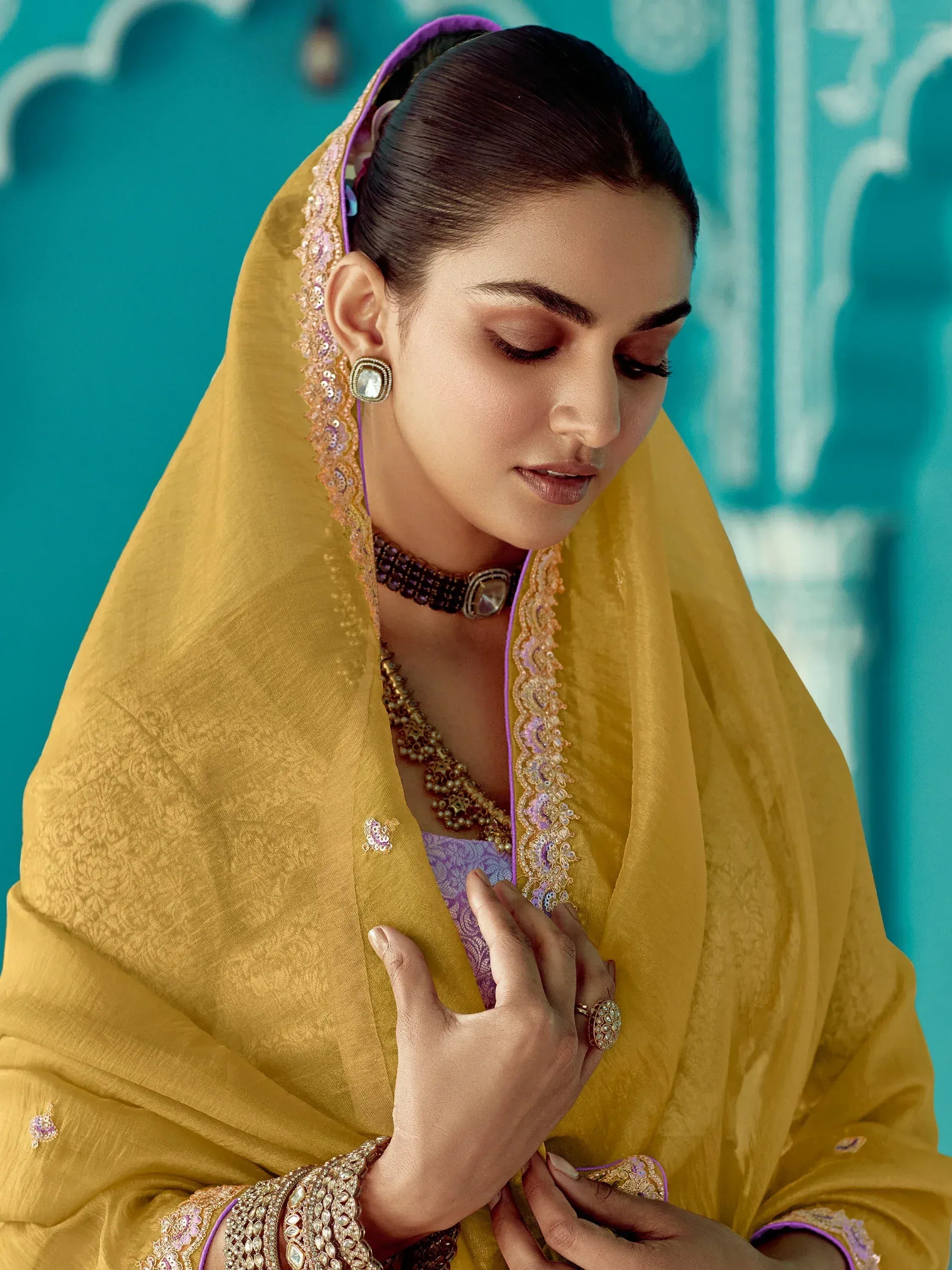 Elegant woman in yellow ethnic wear with purple accents, traditional jewelry, and embroidered dupatta against teal backdrop