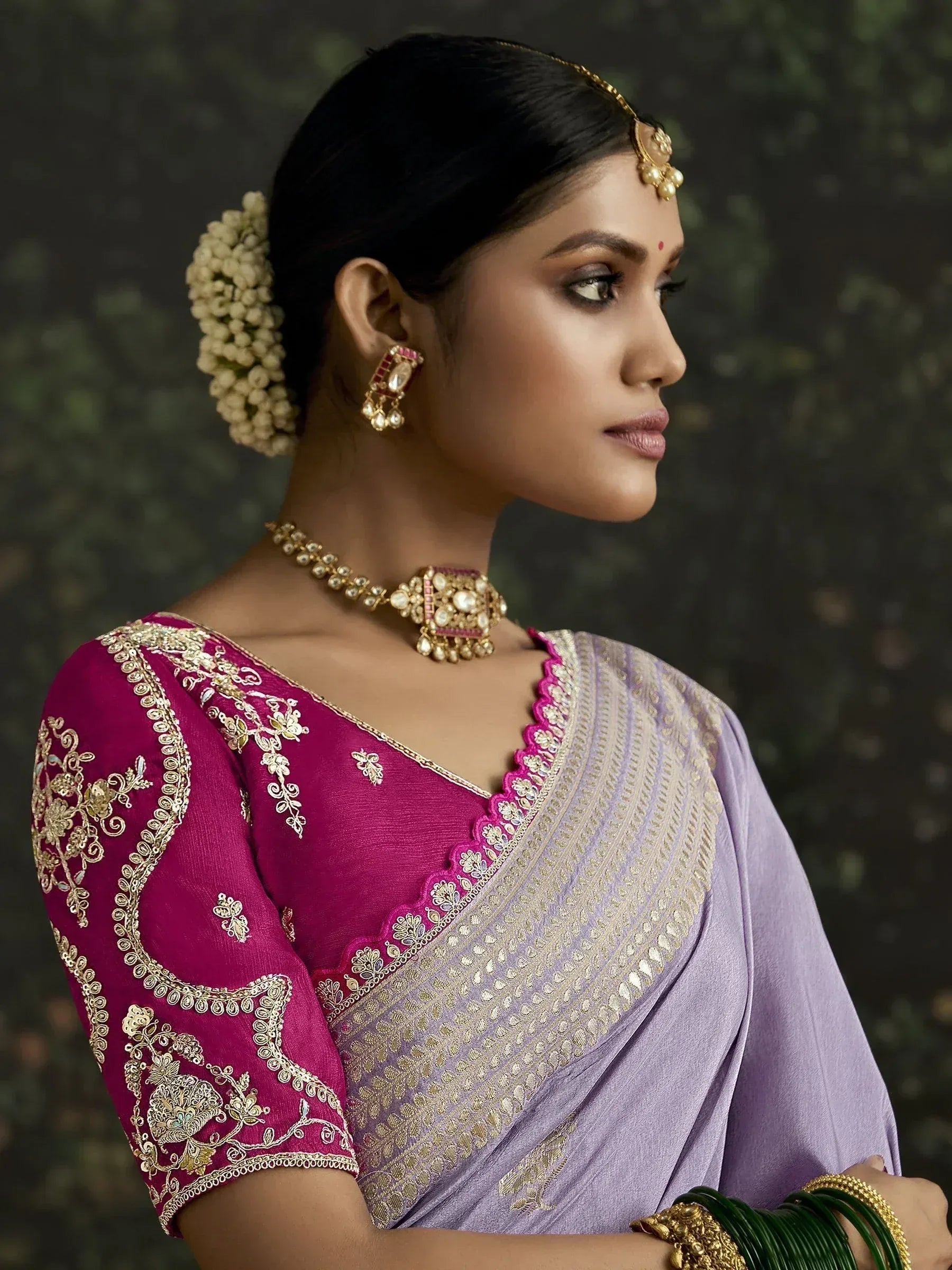 Elegant Indian woman in purple saree with pink embroidered blouse, gold jewelry, and floral hair accessory.