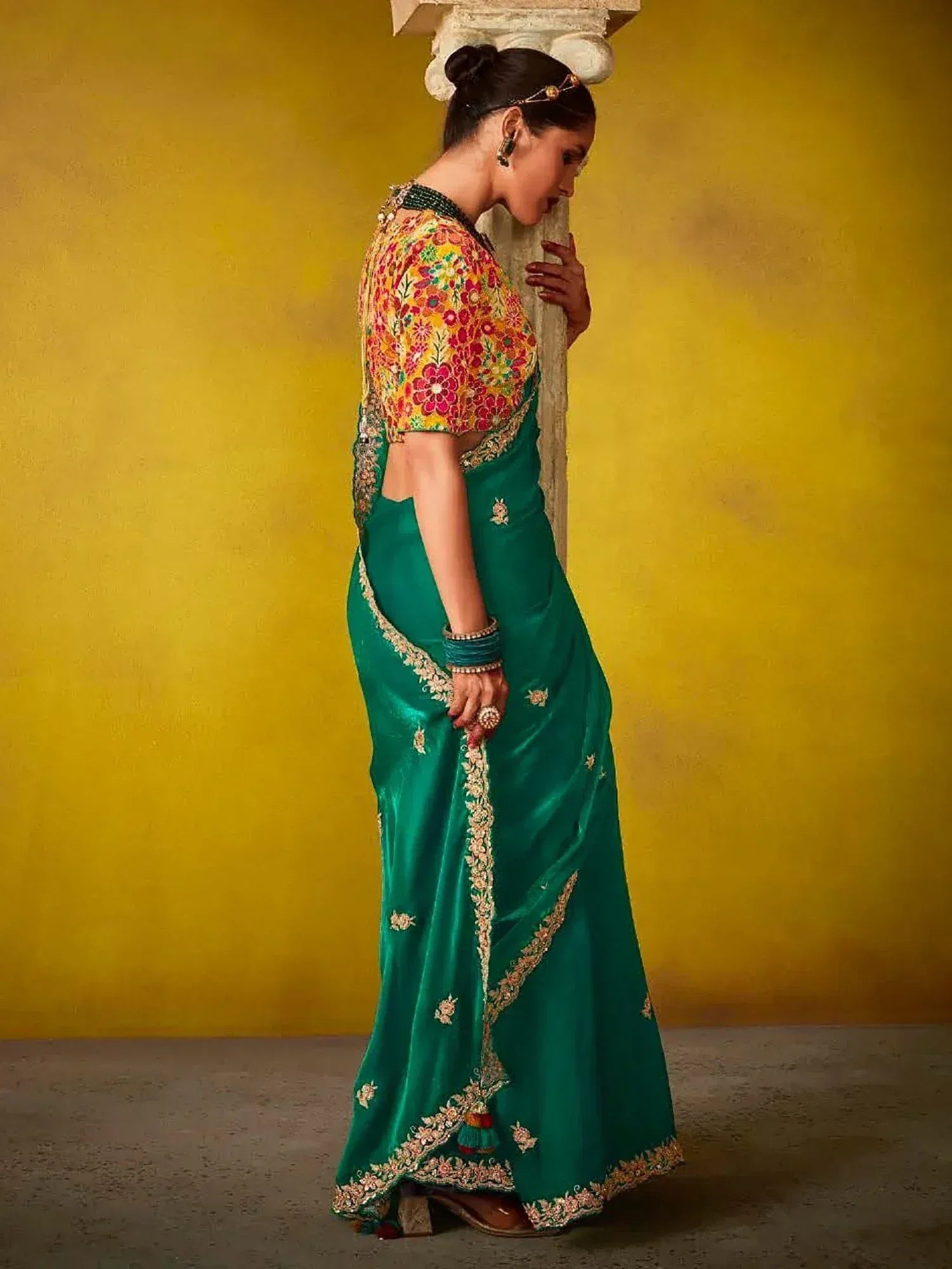 Emerald green saree with floral blouse, gold embroidery, and traditional Indian jewelry against yellow backdrop