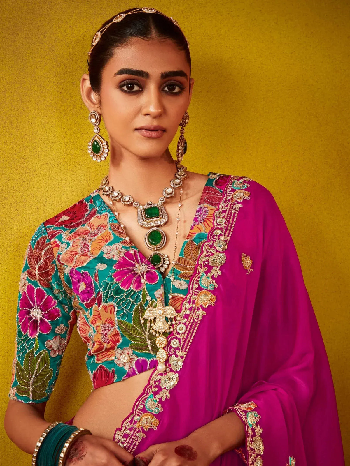 Woman in vibrant floral embroidered Indian ethnic wear with green gemstone jewelry against gold backdrop