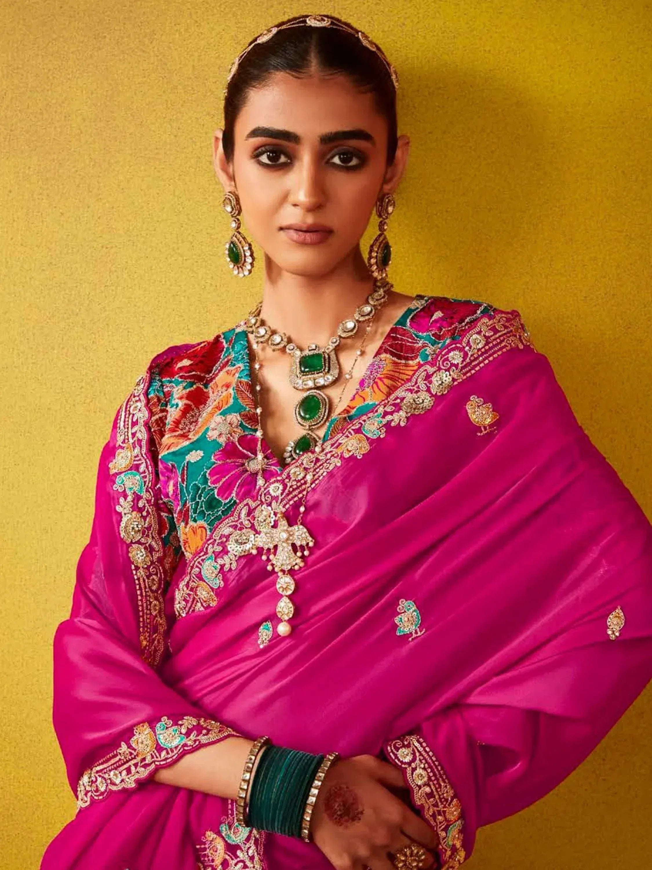 Woman in vibrant pink embroidered Indian ethnic wear with green jewelry against gold background