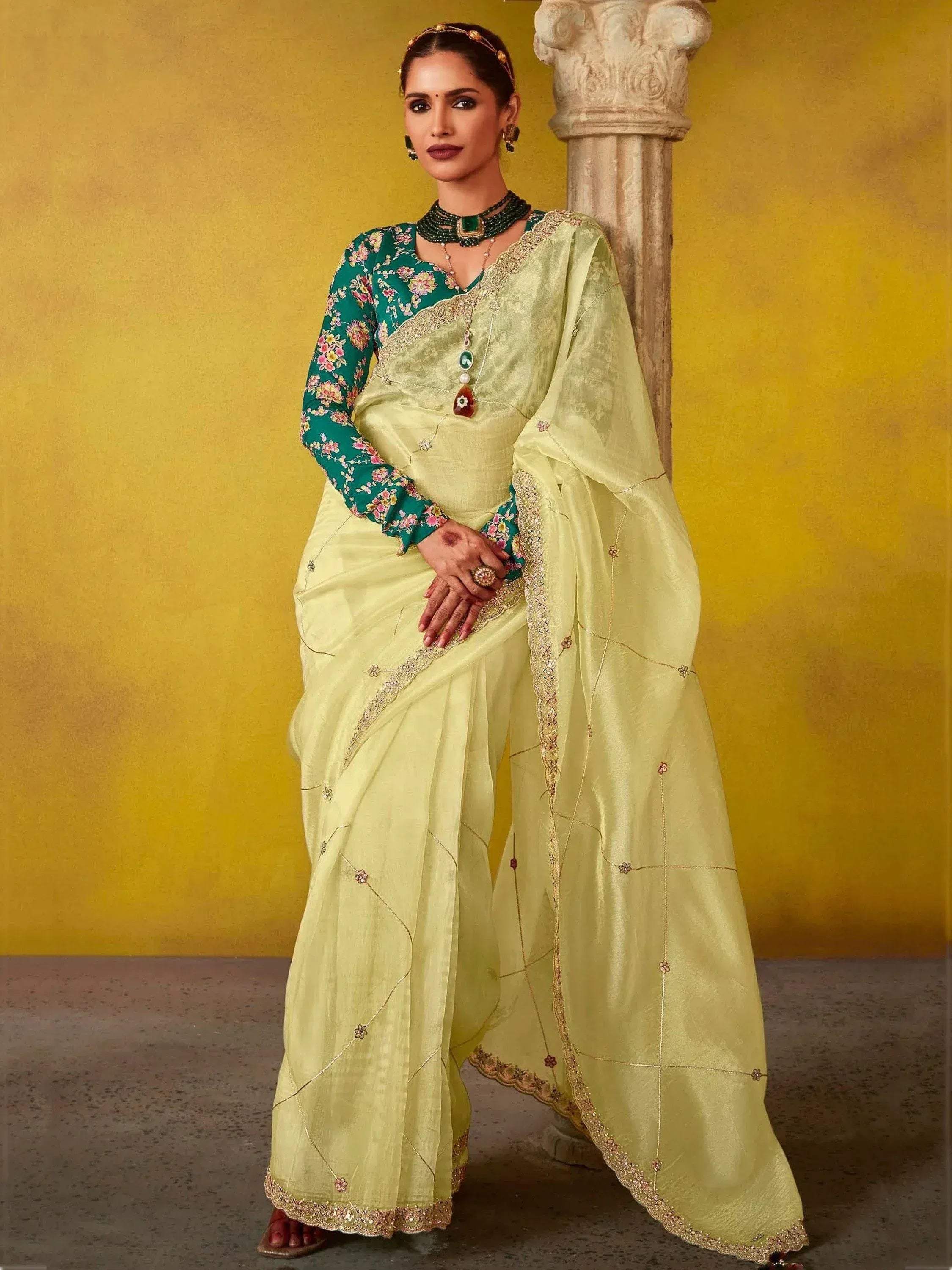 Woman in light green saree with floral blouse, gold embroidery, and traditional jewelry against yellow backdrop