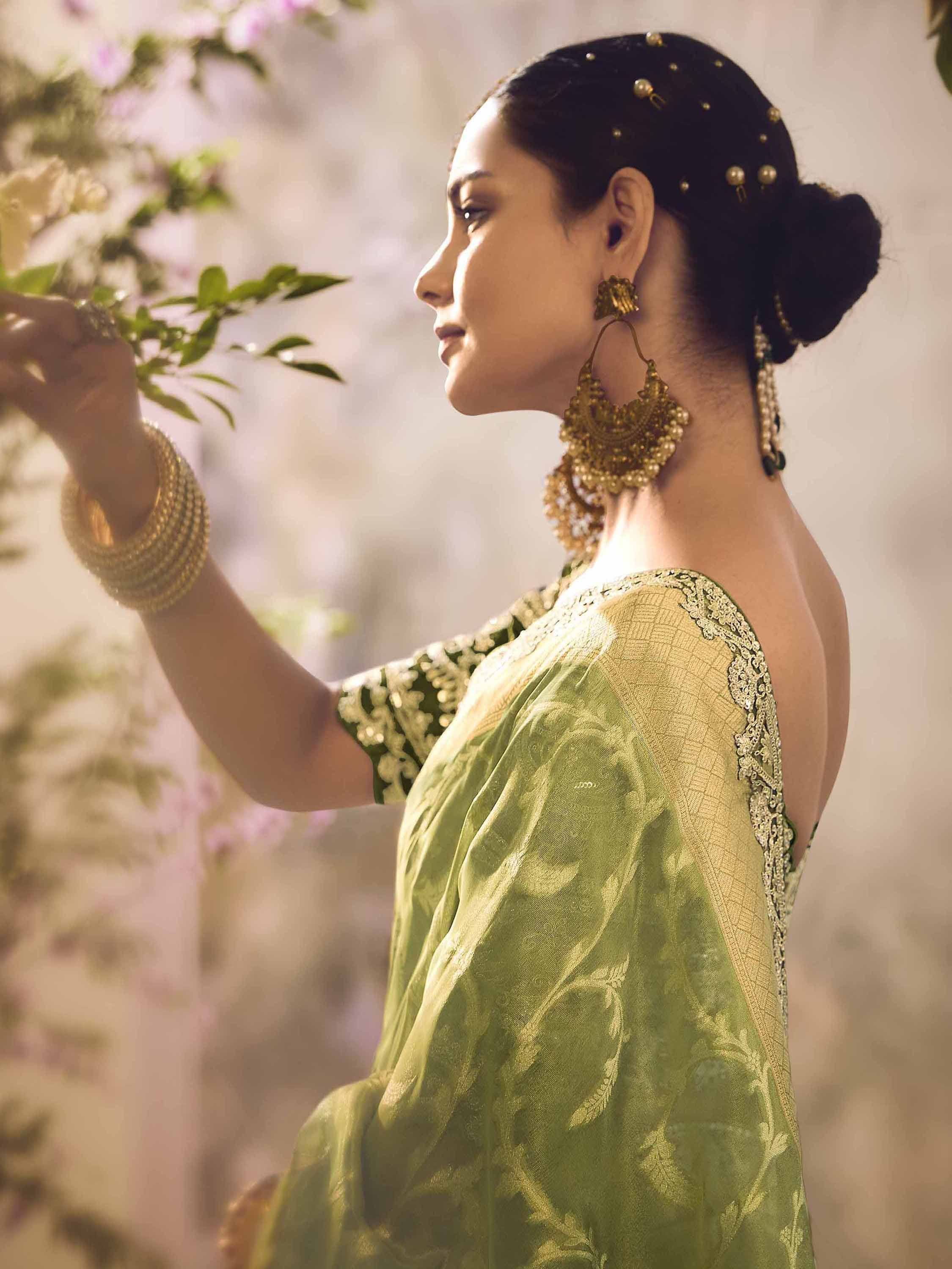 Elegant woman in green designer saree with gold embroidery, traditional jewelry, and floral backdrop