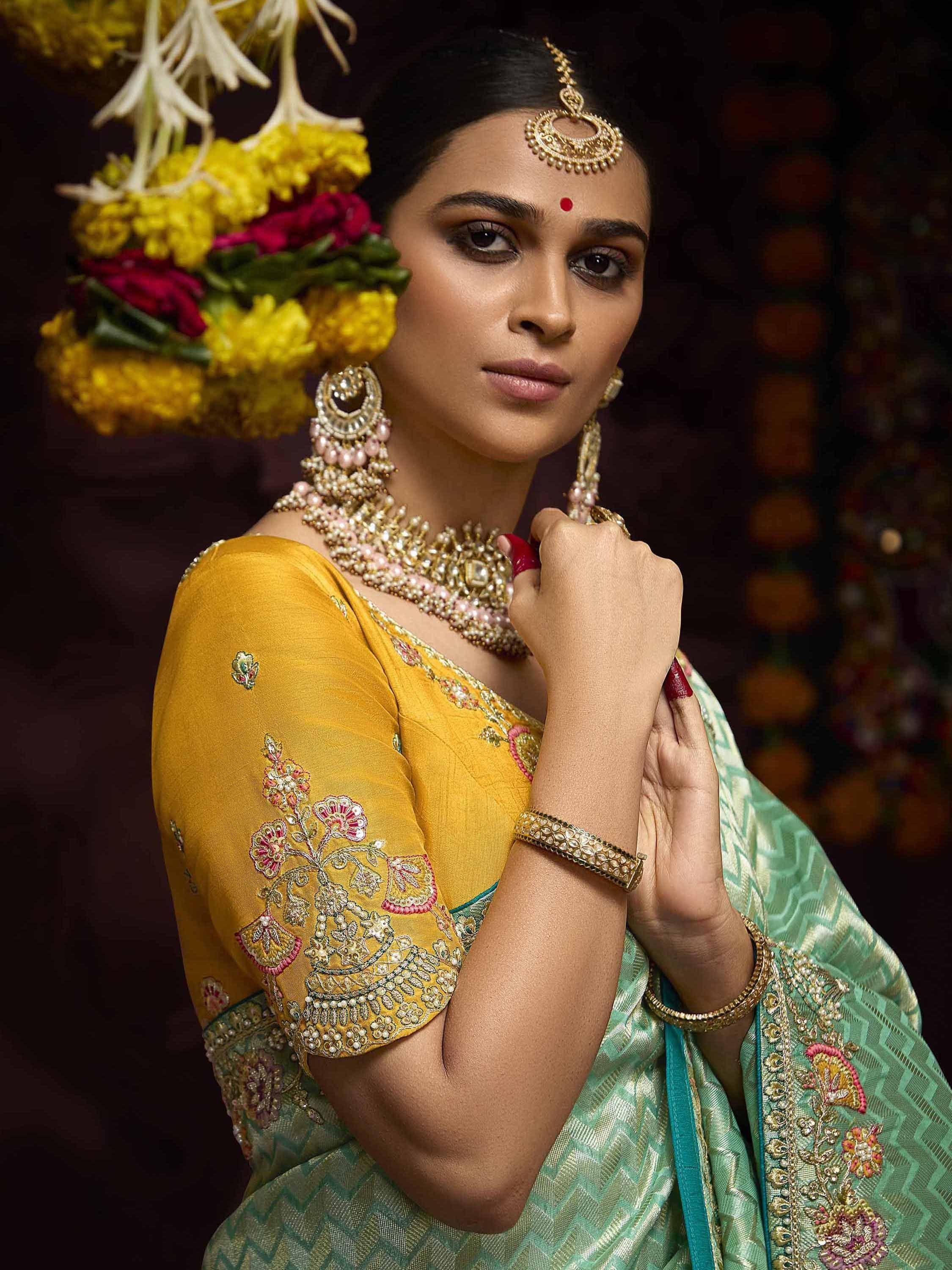 Woman in embroidered yellow blouse and green saree with gold jewelry and floral backdrop