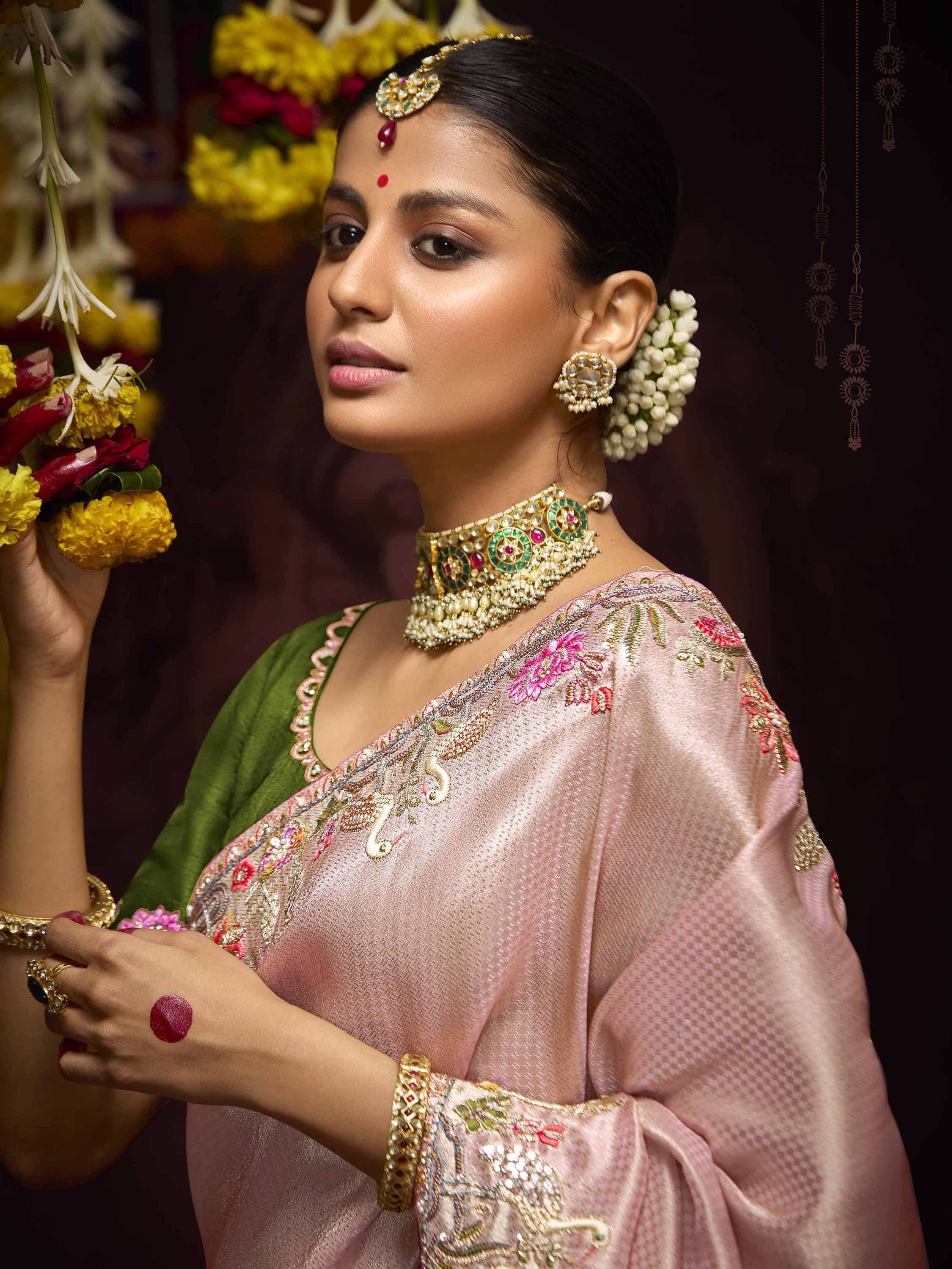 Woman in pink embroidered saree with gold jewelry and floral hair accessory, holding marigold garland