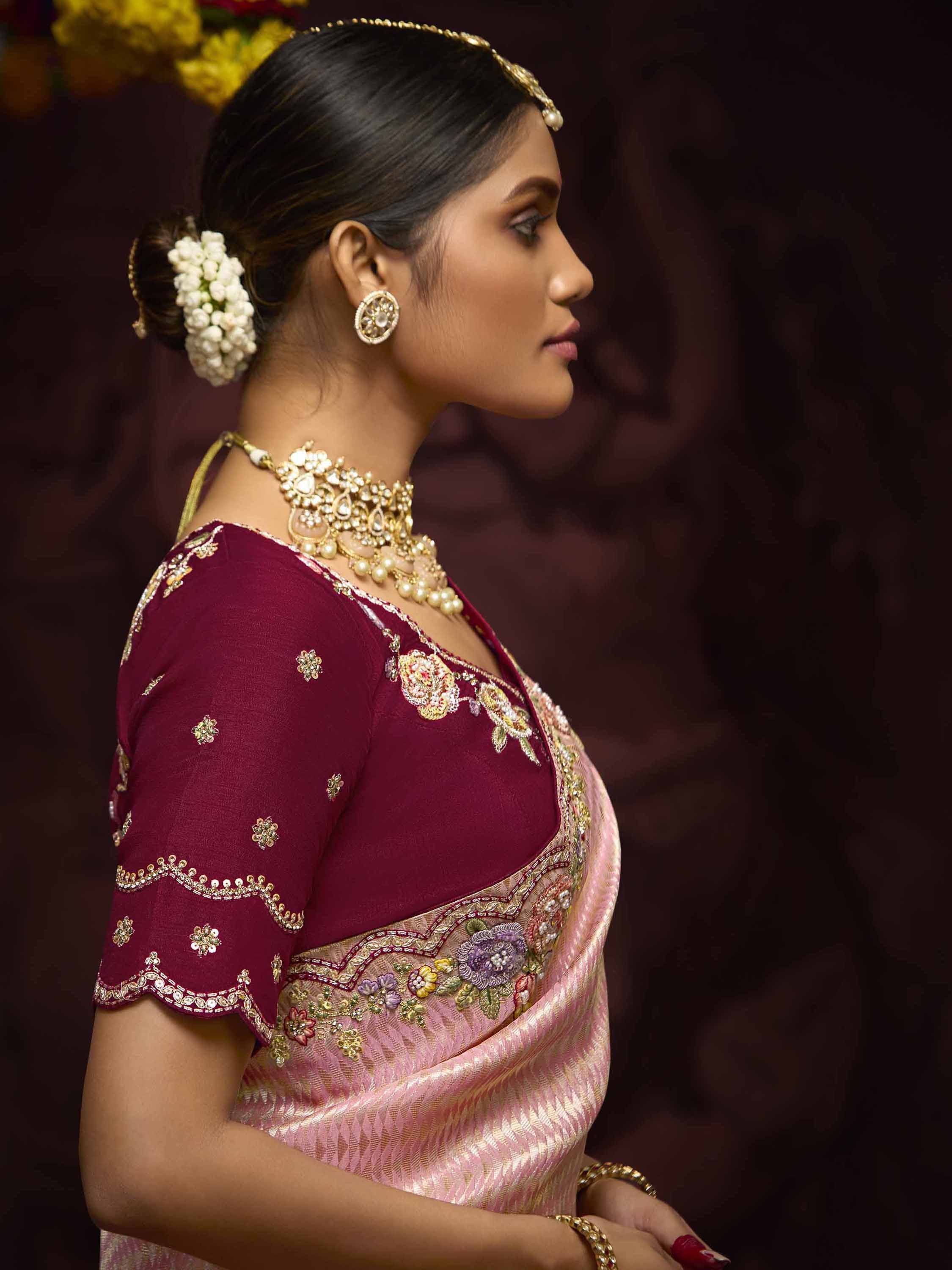 Woman in maroon embroidered blouse and pink saree with gold jewelry, floral hair accessory
