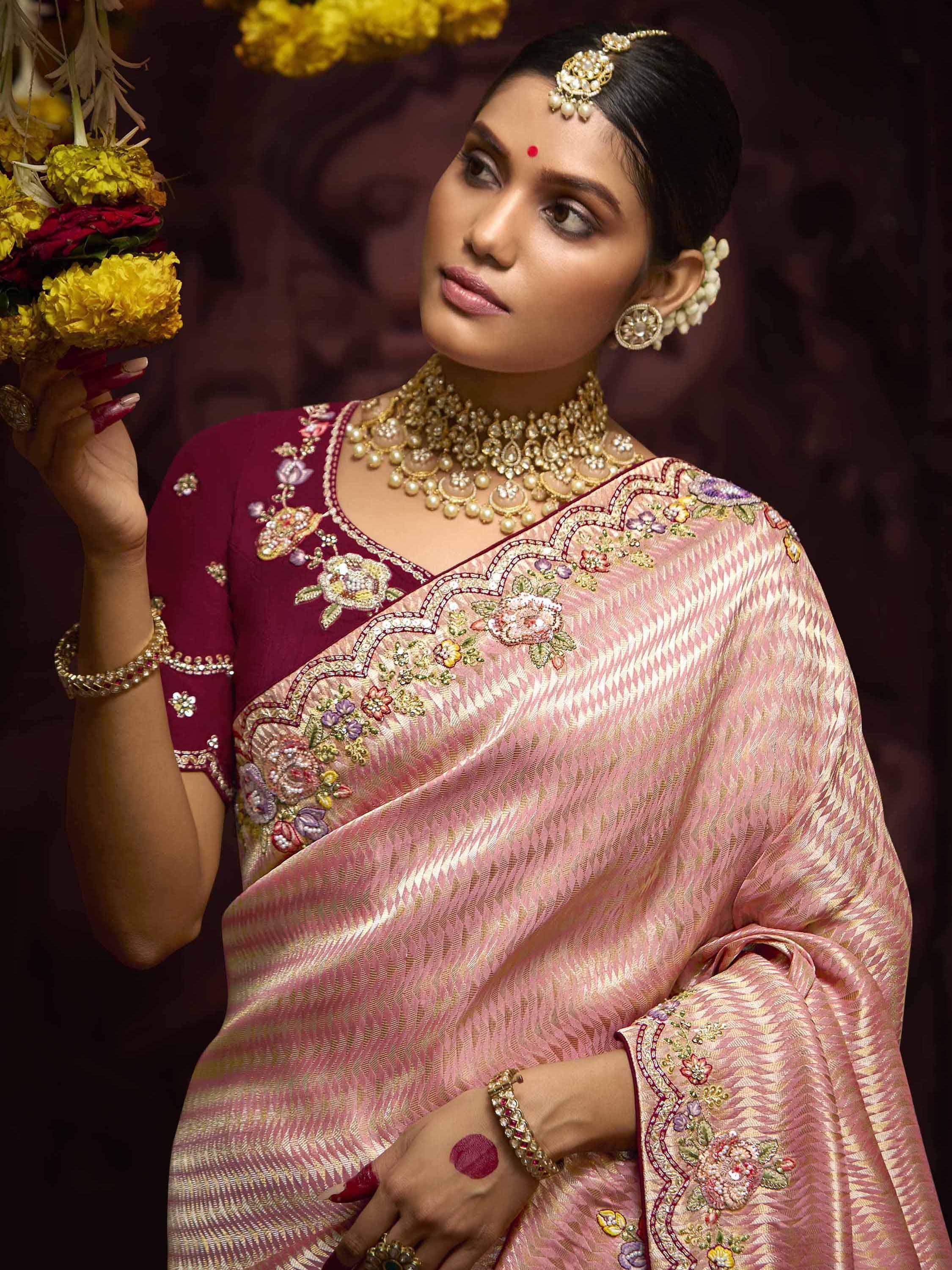 Elegant woman in pink and maroon embroidered saree with floral motifs, traditional jewelry, and yellow flowers.