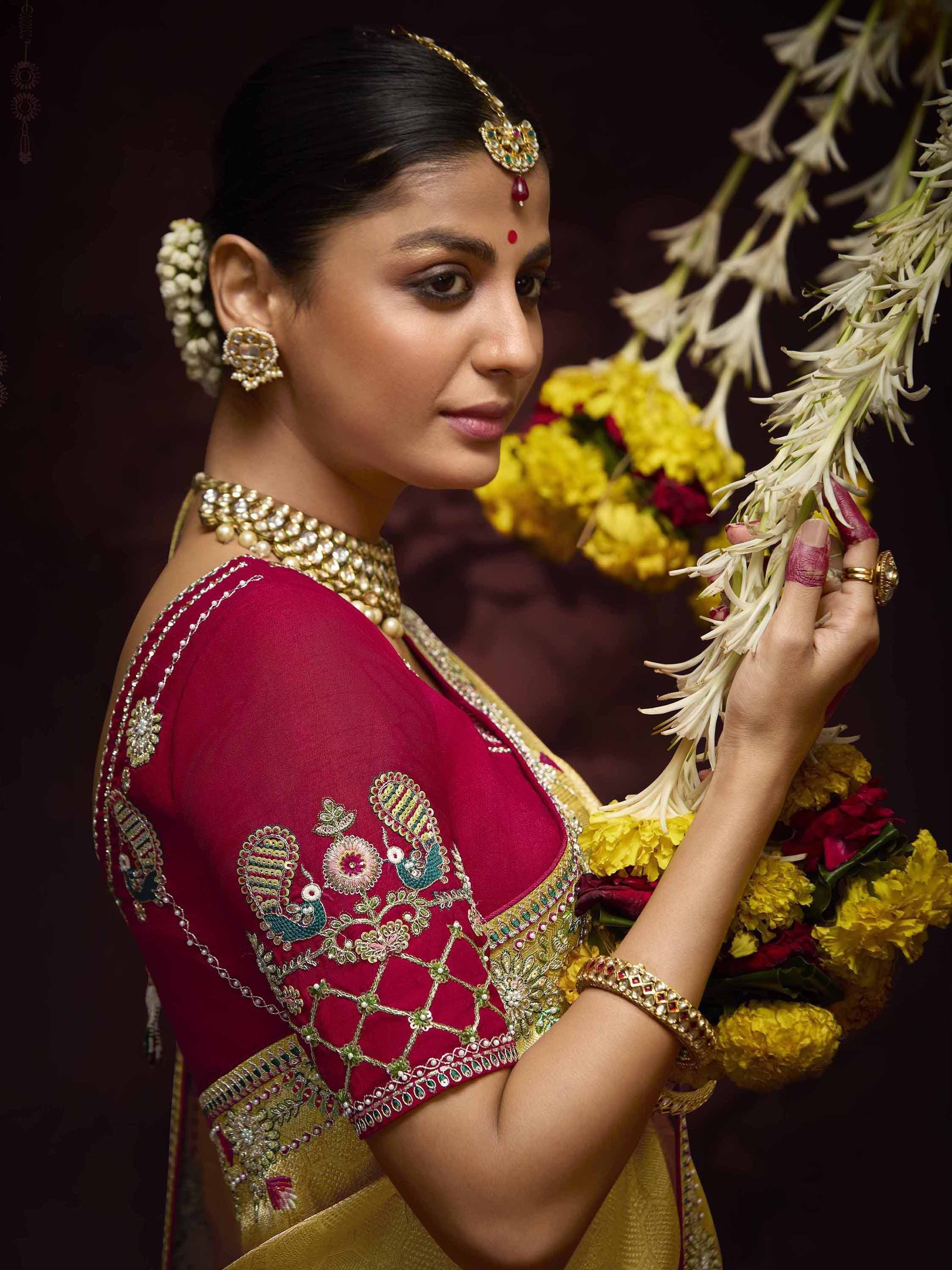 Woman in maroon embroidered blouse with gold saree, traditional Indian jewelry, and floral garland