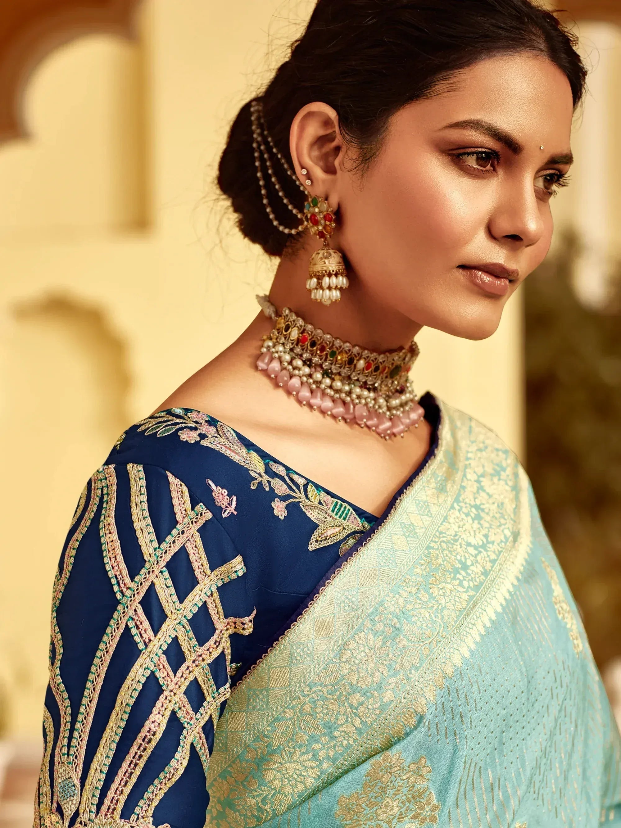 Elegant woman in embroidered blue blouse and light blue saree with gold detailing, adorned with traditional jewelry.