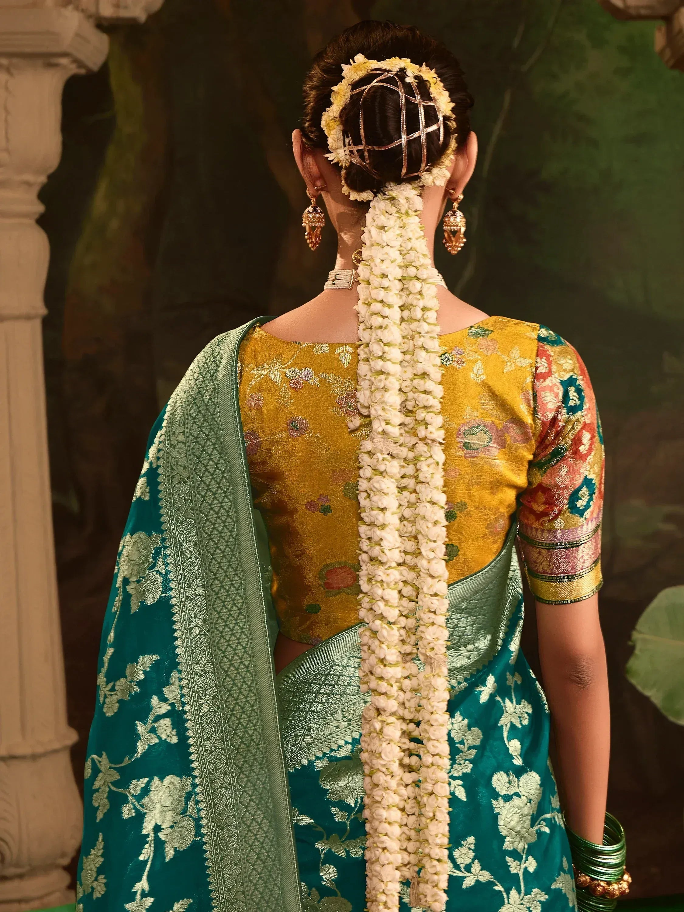 Woman in teal floral saree with yellow blouse, jasmine gajra, and gold jewelry, showcasing traditional Indian ethnic wear.