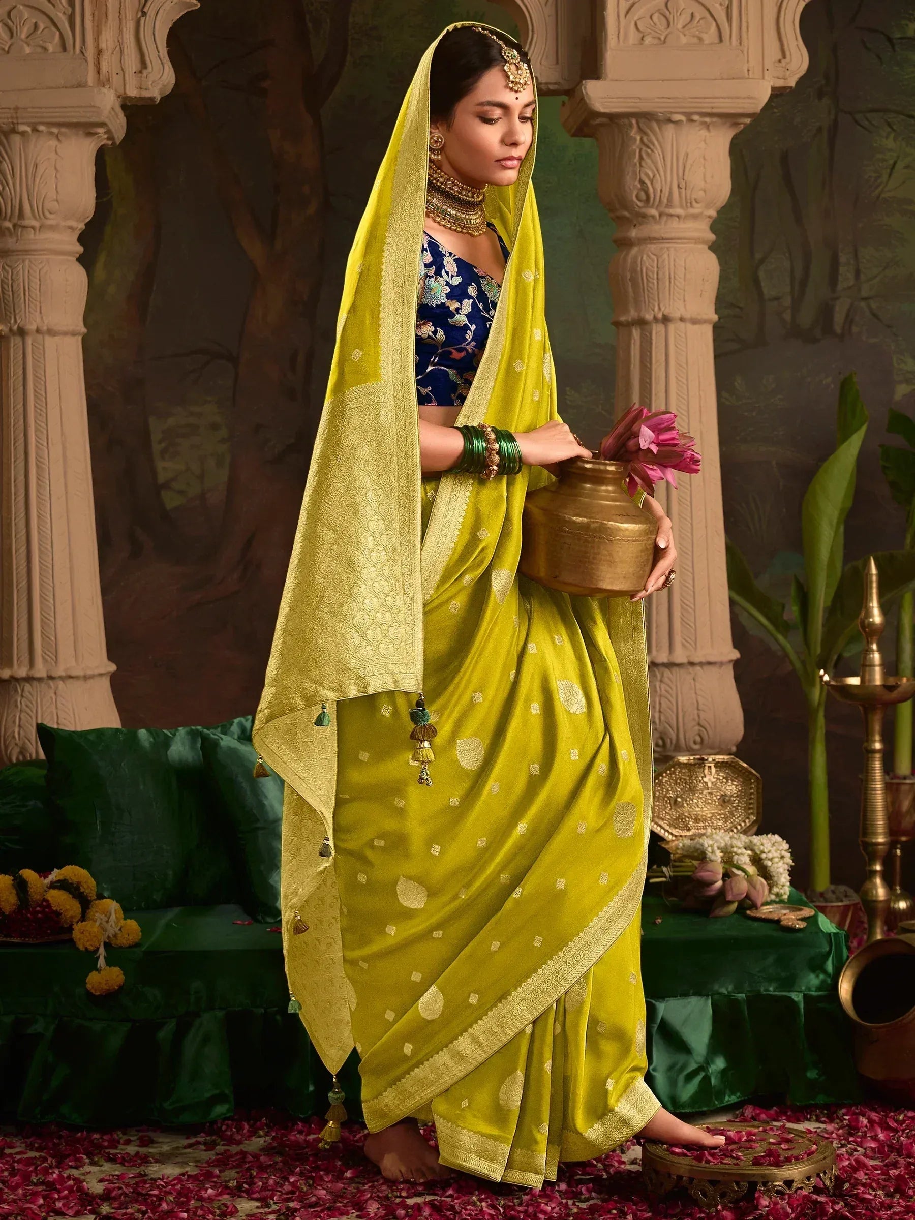 Elegant woman in vibrant yellow saree with gold embroidery, holding a brass pot, surrounded by floral decor.