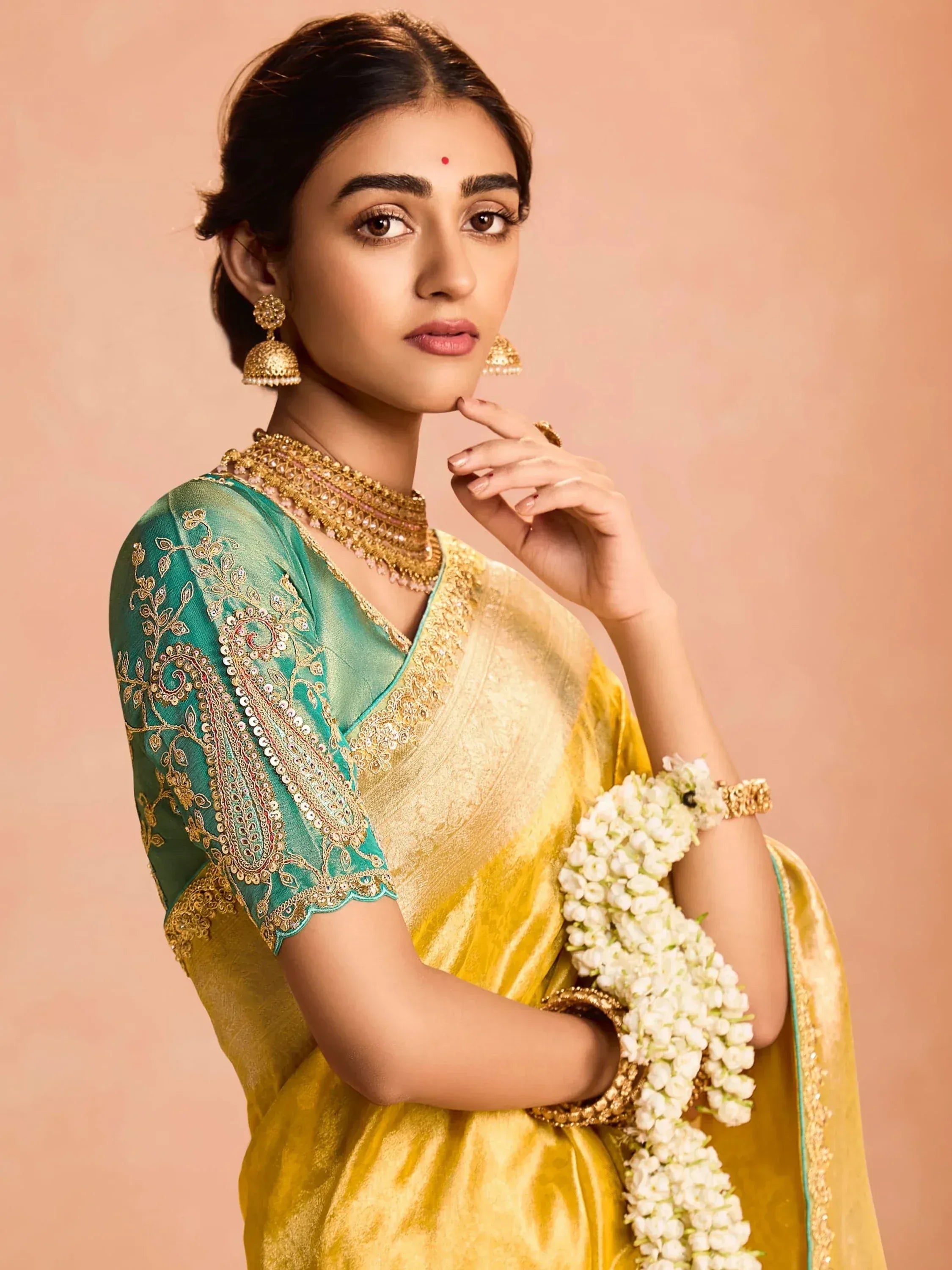 Elegant woman in golden saree with green embroidered blouse, traditional jewelry, and floral garland