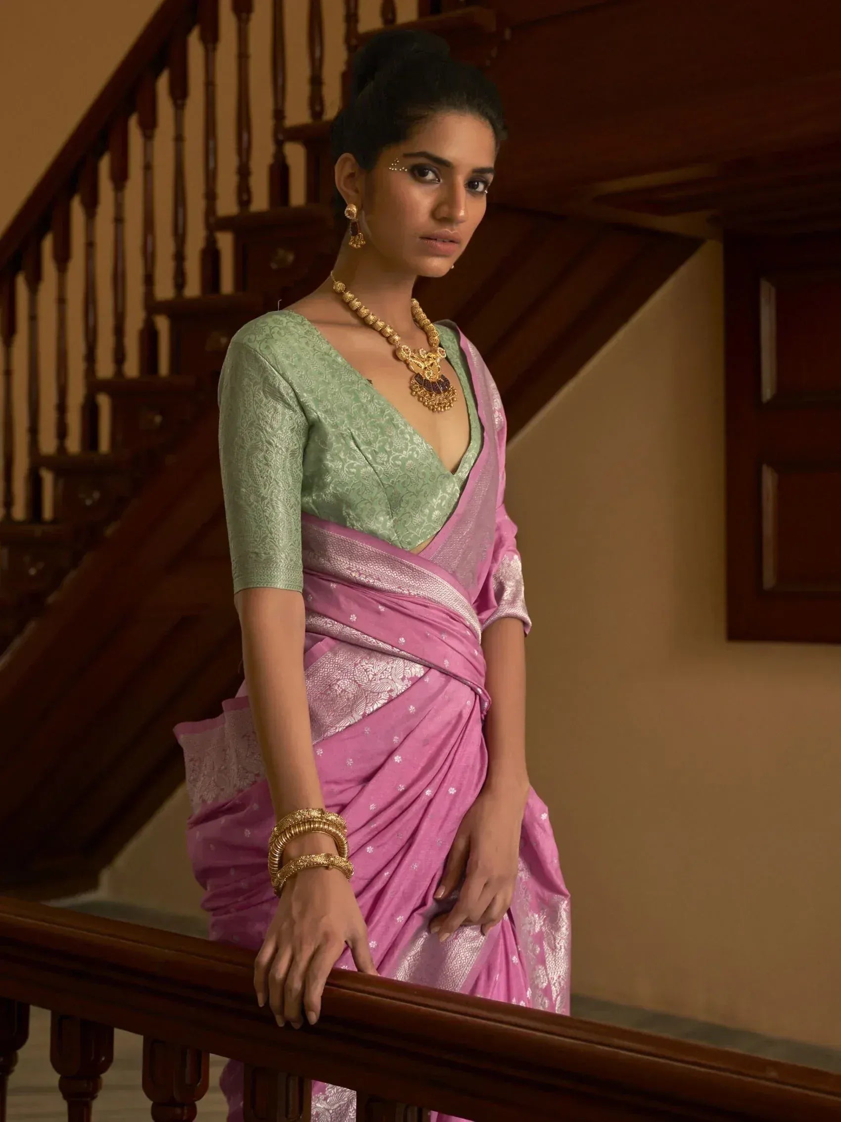 Woman in pink saree with green blouse, gold jewelry, standing by wooden staircase