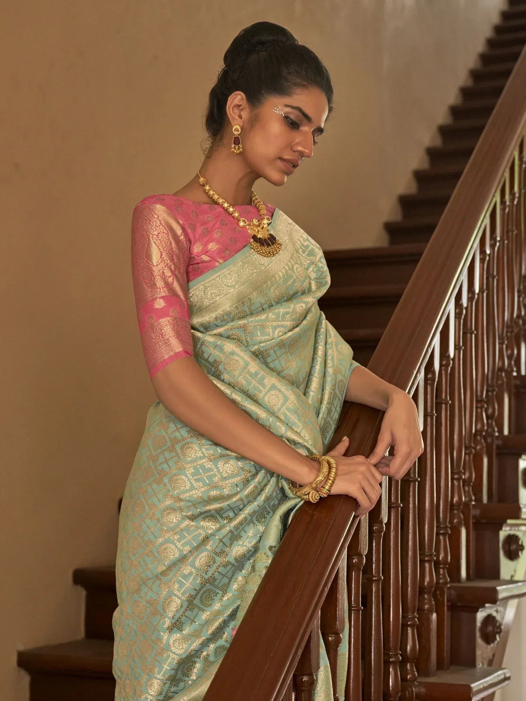 Woman in mint green and pink designer saree with gold embroidery, traditional jewelry, leaning on wooden staircase