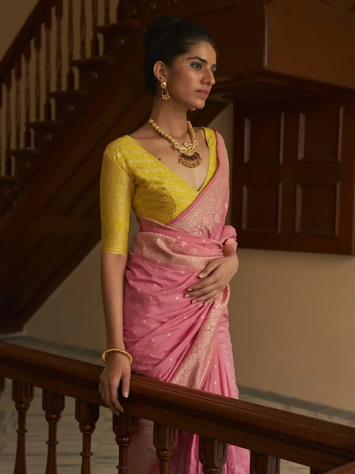 Woman in pink saree with yellow blouse, gold jewelry, standing by wooden staircase