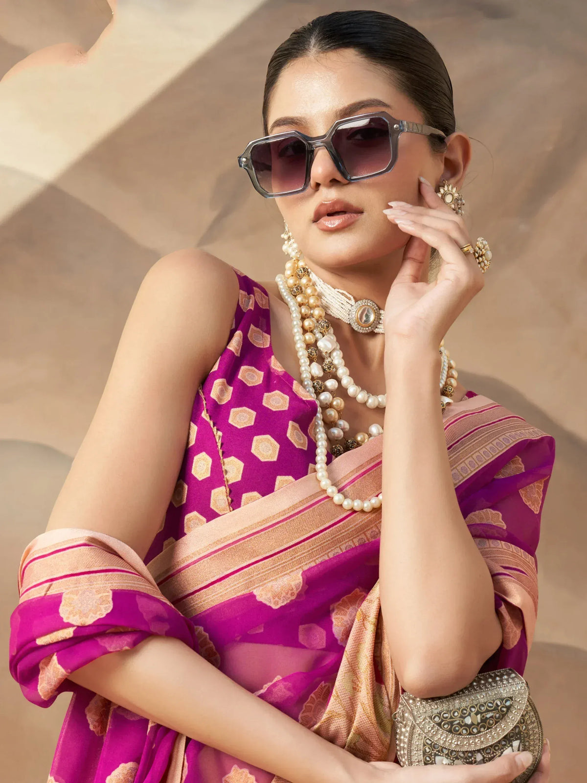 Woman in purple printed saree with gold border, pearl jewelry, and statement sunglasses, holding embellished clutch.