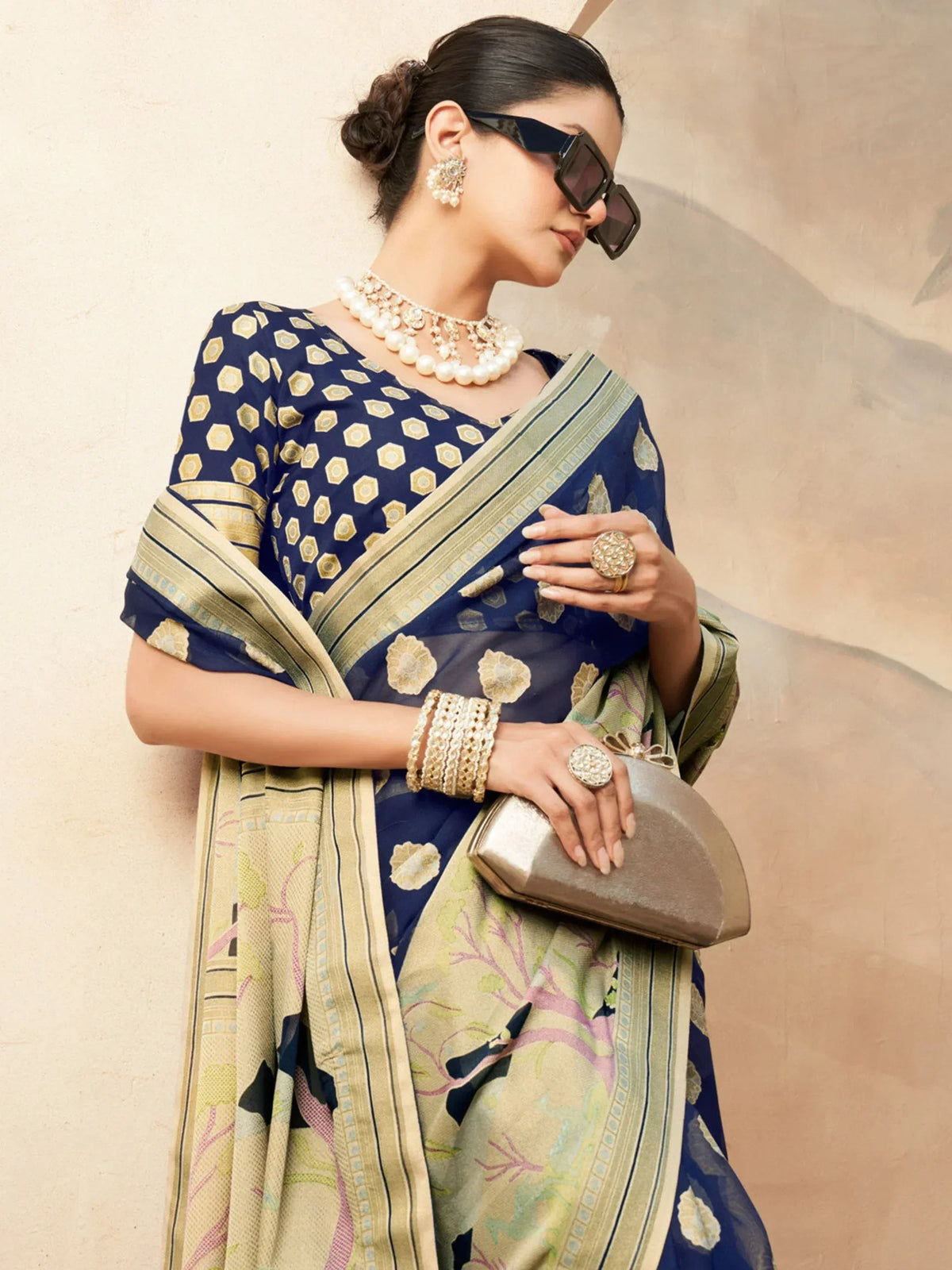 Woman in navy blue printed saree with gold border, pearl jewelry, and sunglasses, holding clutch