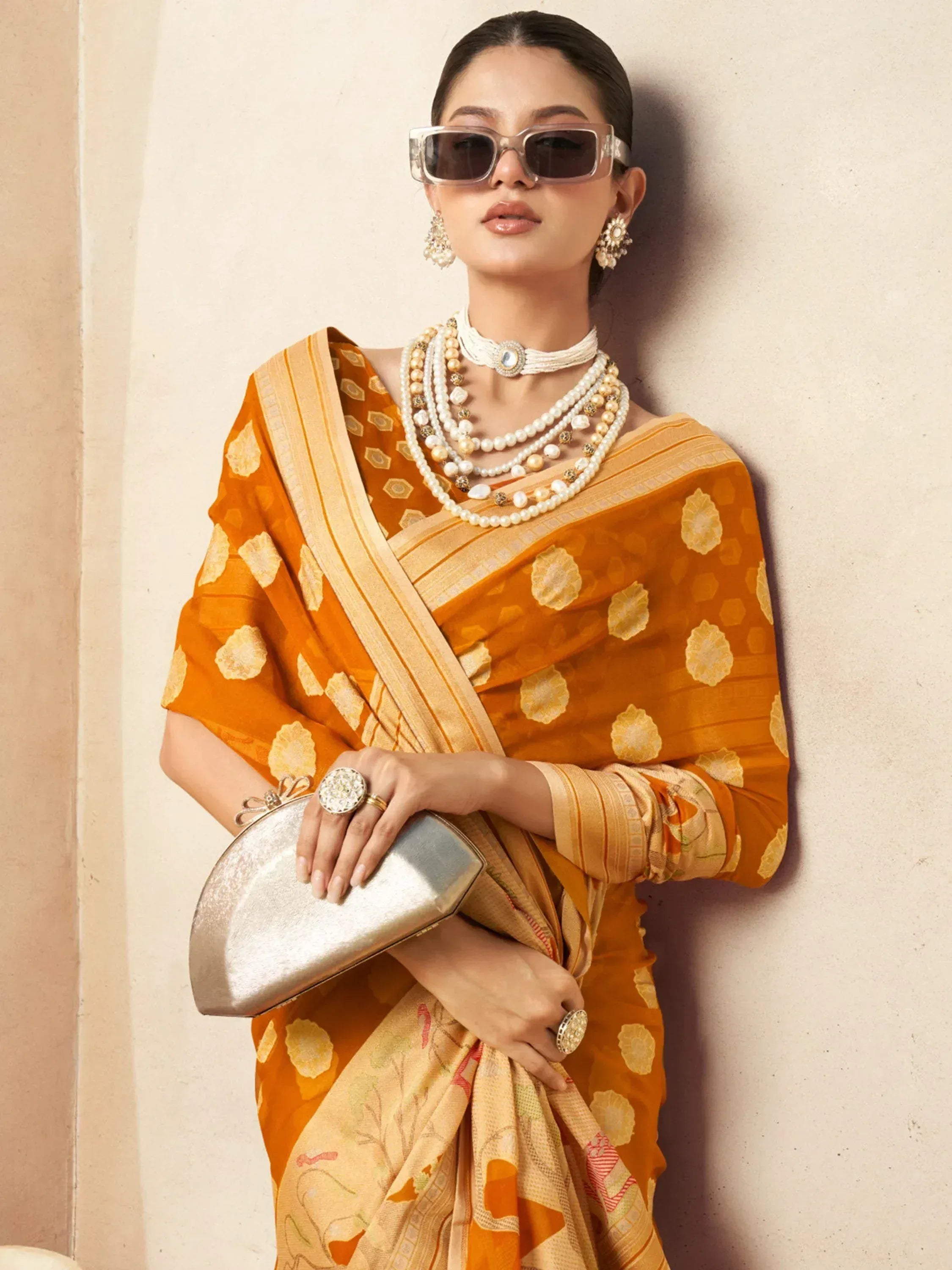 Woman in vibrant orange printed saree with pearl jewelry and sunglasses, holding metallic clutch